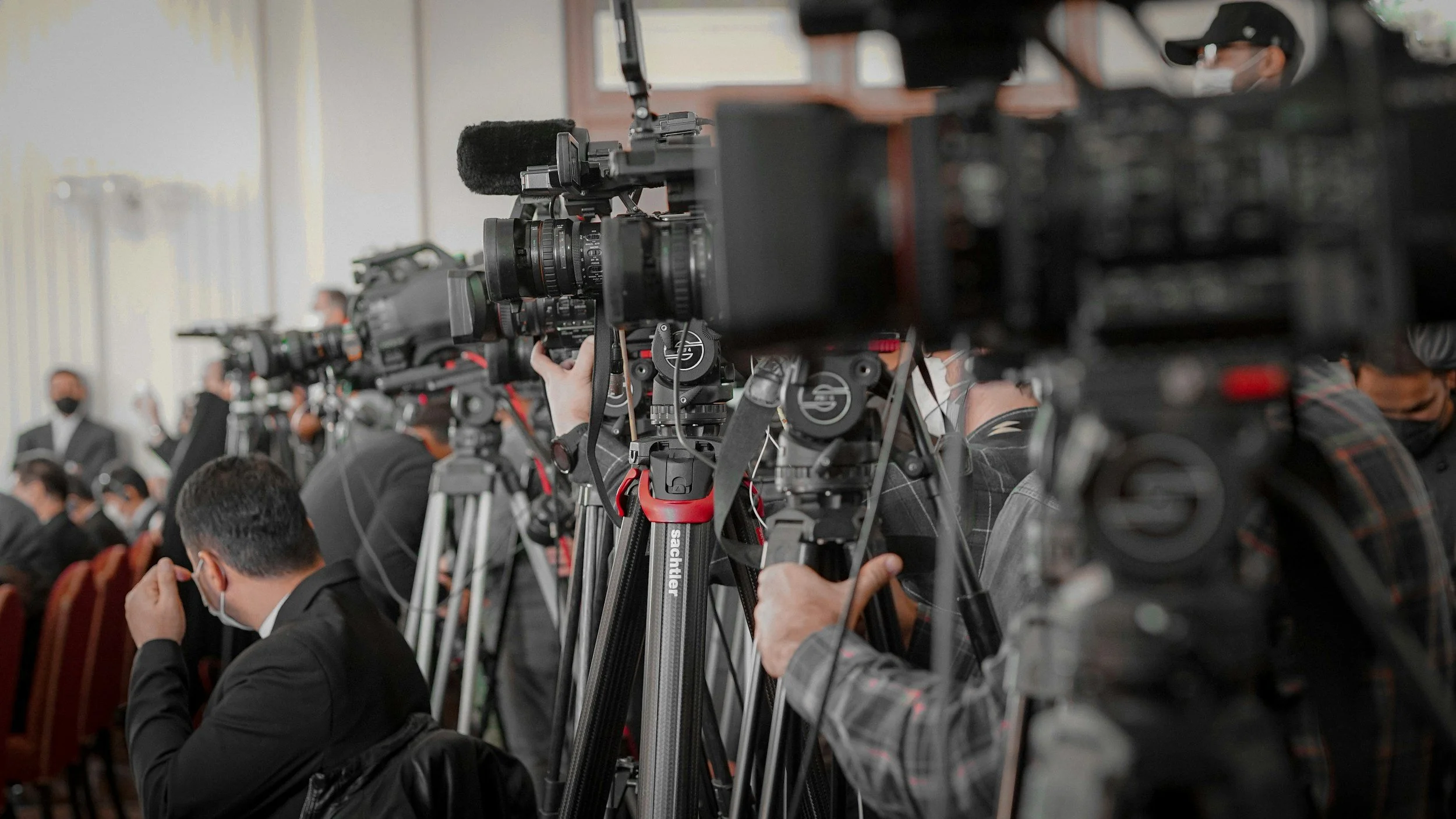 Several TV cameras on tripods and photographers seated in an indoor event, all wearing masks.