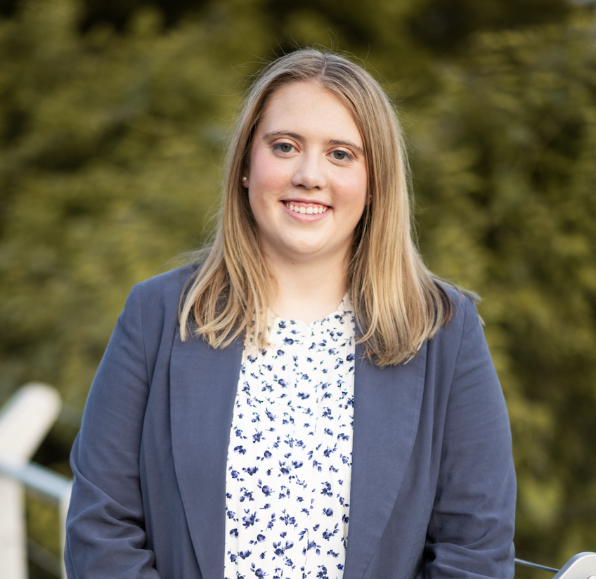 A woman with shoulder-length blonde hair, wearing a navy blazer over a white blouse with blue floral pattern, smiling outdoors with blurred green trees in the background.