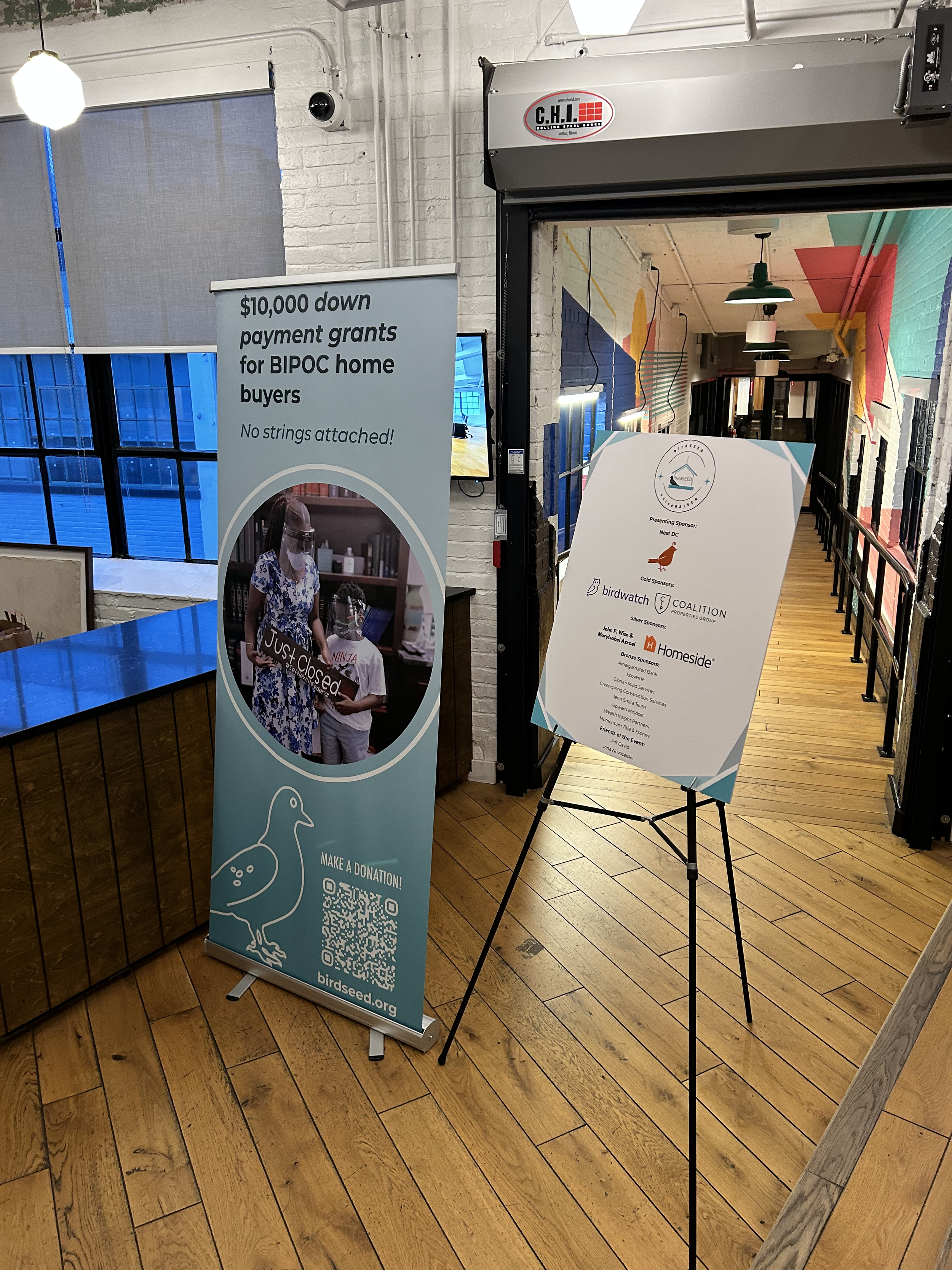 Indoor space with a wooden floor, colorful abstract wall art, and signage promoting a birdseed donation event. One sign advertises a $10,000 down payment grant for BIPOC home buyers with a QR code and an image of a woman with a child holding a sign that says 'Just Closed.' Another easel sign lists sponsors of the event.