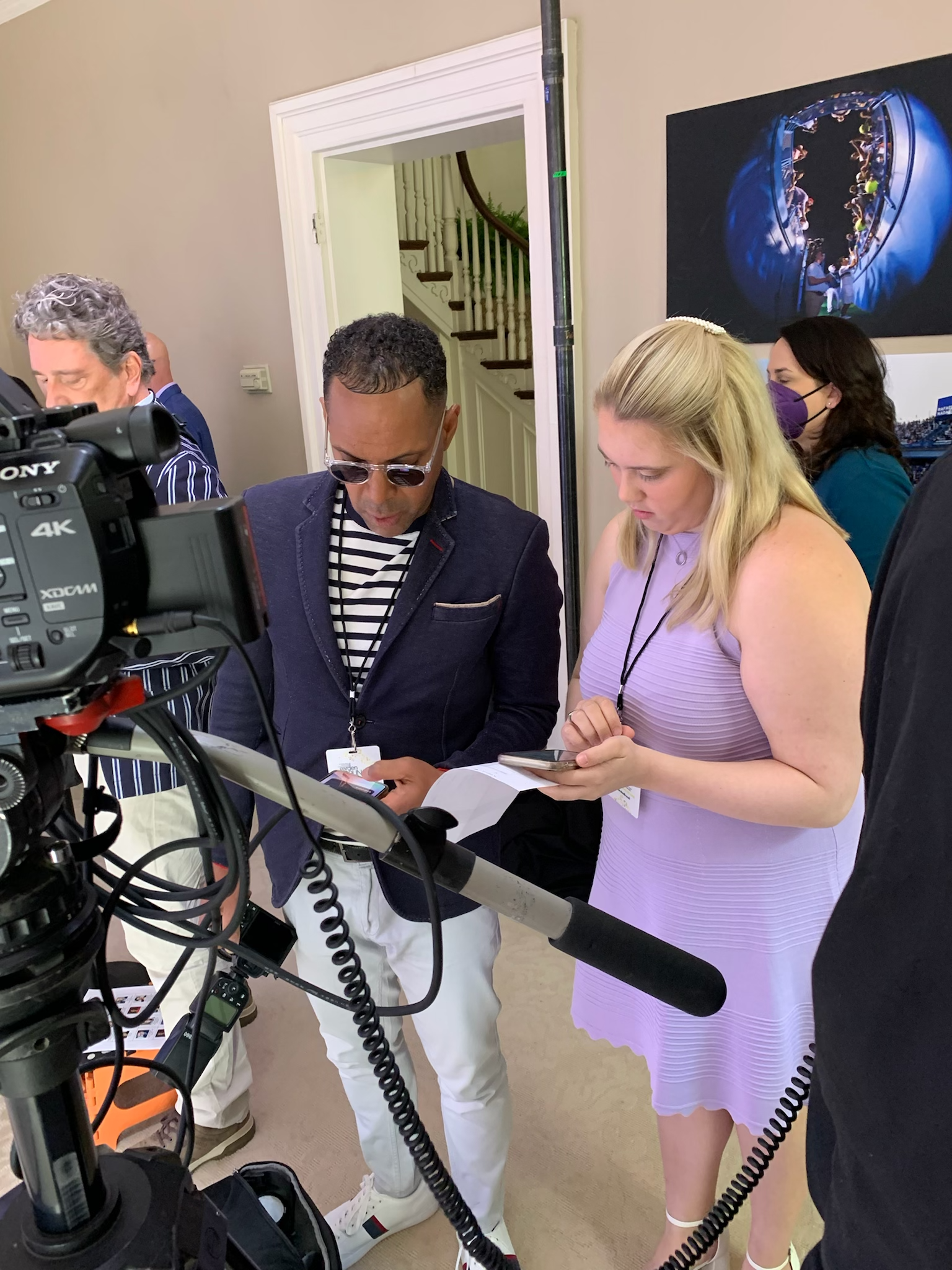 Group of people working with a professional video camera in an indoor setting, some looking at their phones, with framed photos on the wall.