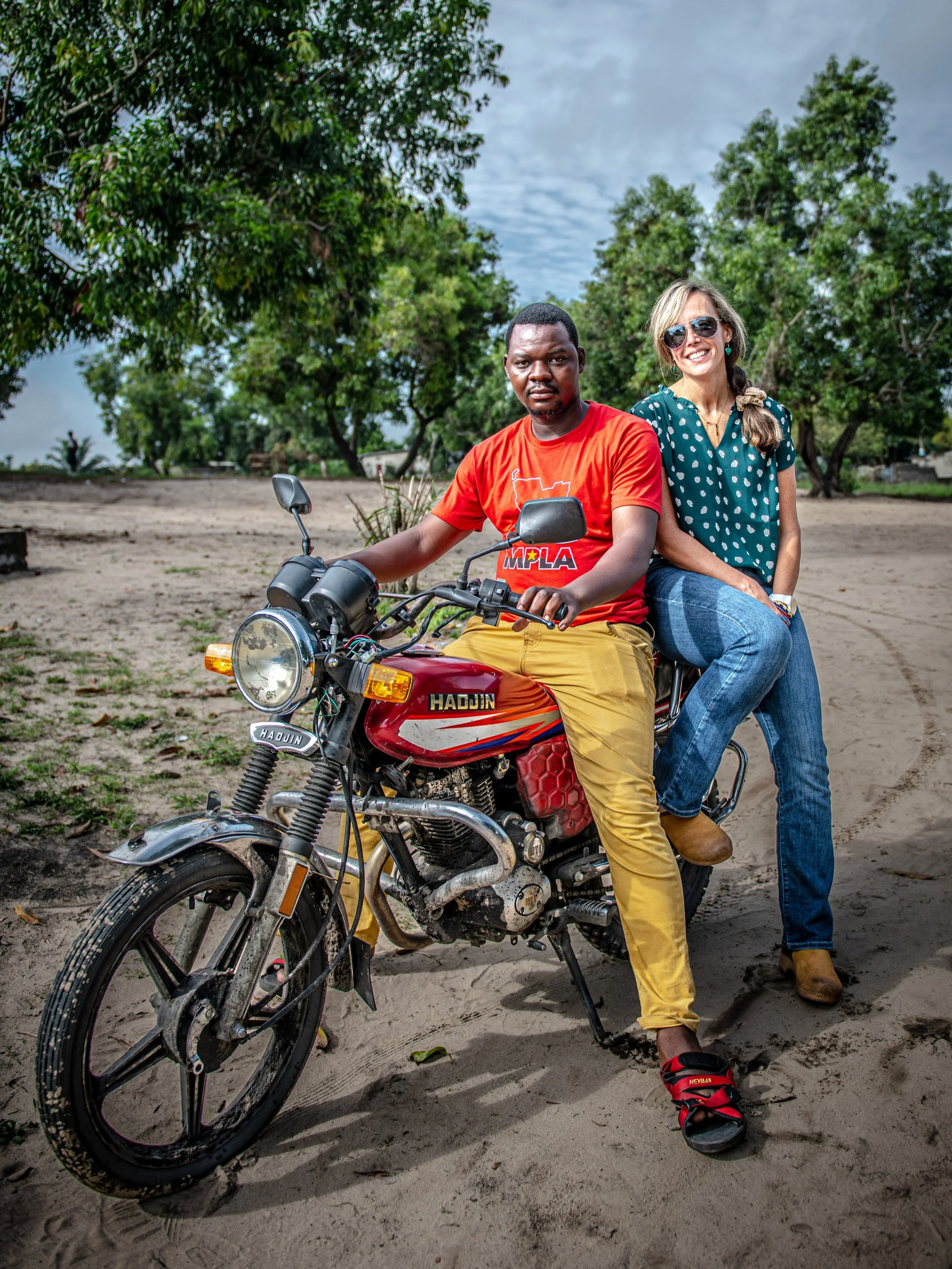 Two people sitting on a motorcycle in an outdoor setting with trees in the background. The person in front is wearing a red T-shirt and yellow pants, while the person behind is wearing sunglasses and casual clothing. The motorcycle is parked on sandy ground.
