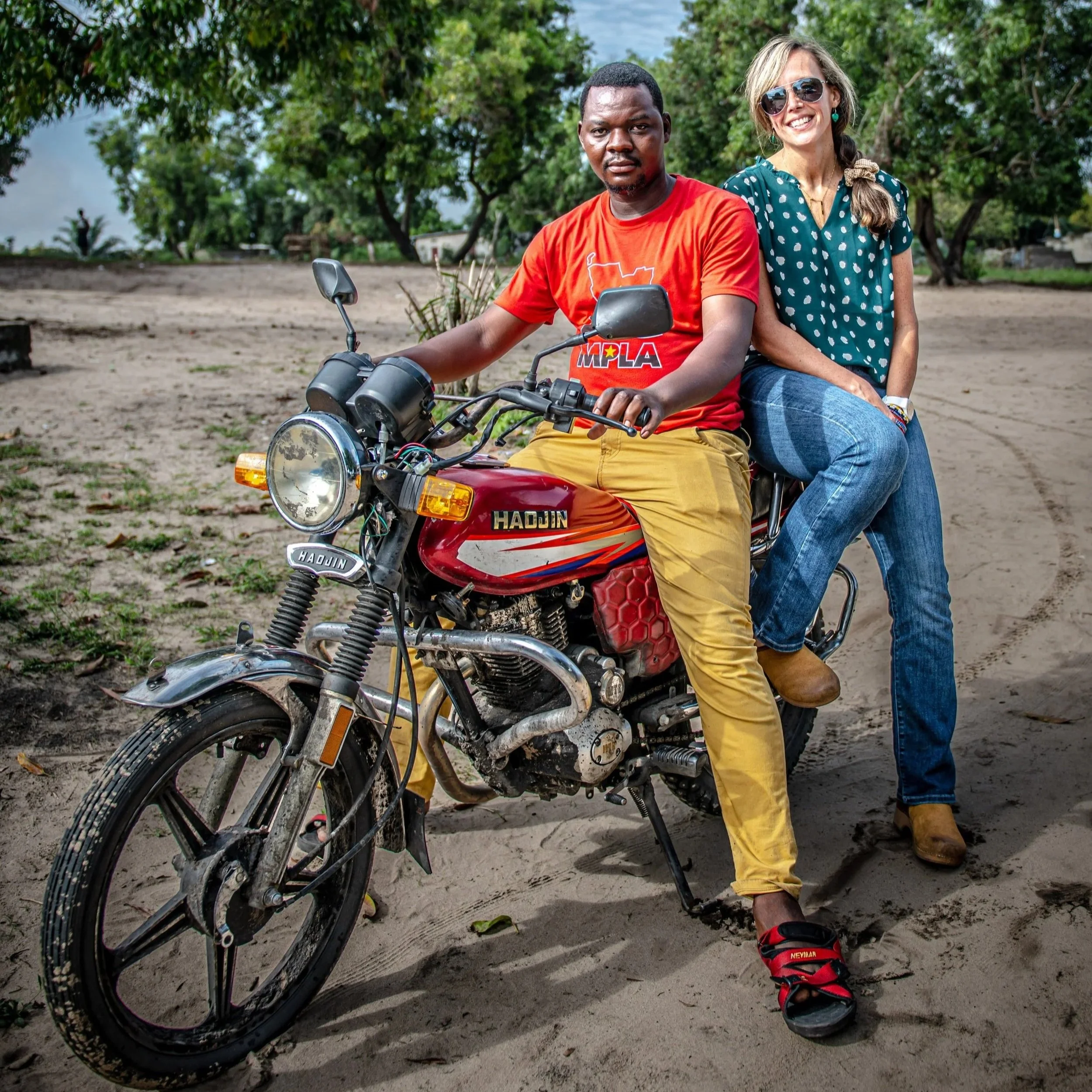 A man and woman sitting on a red motorcycle outdoors on a dirt path with green trees in the background.