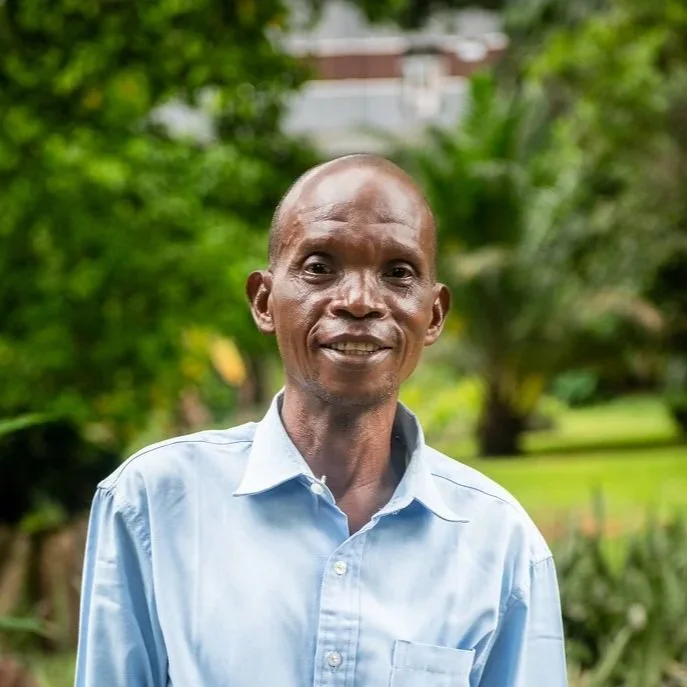 Portrait of an elderly man with a bald head, smiling, wearing a light blue button-up shirt in a green outdoor setting.