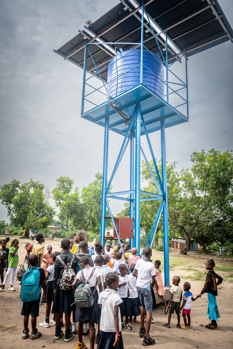 A group of children and adults gathered around a water tower with solar panels on top in an outdoor area, with trees and a cloudy sky in the background.