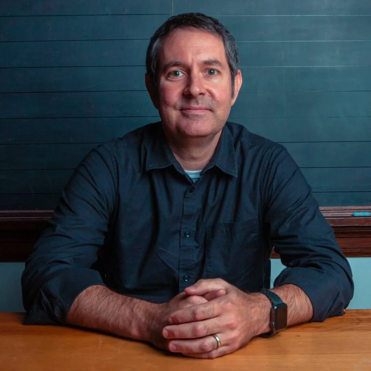 A man with short dark hair and a slight smile, wearing a dark shirt, sits at a wooden table with hands clasped, in front of a dark green wall.