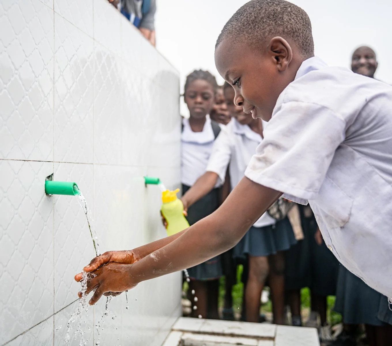 A young boy in a school uniform washing his hands with water at an outdoor handwashing station, with several students observing.