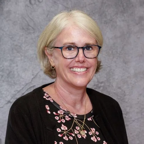 A smiling woman with short blonde hair, glasses, and earrings, wearing a black cardigan over a floral blouse, standing in front of a textured gray wall.