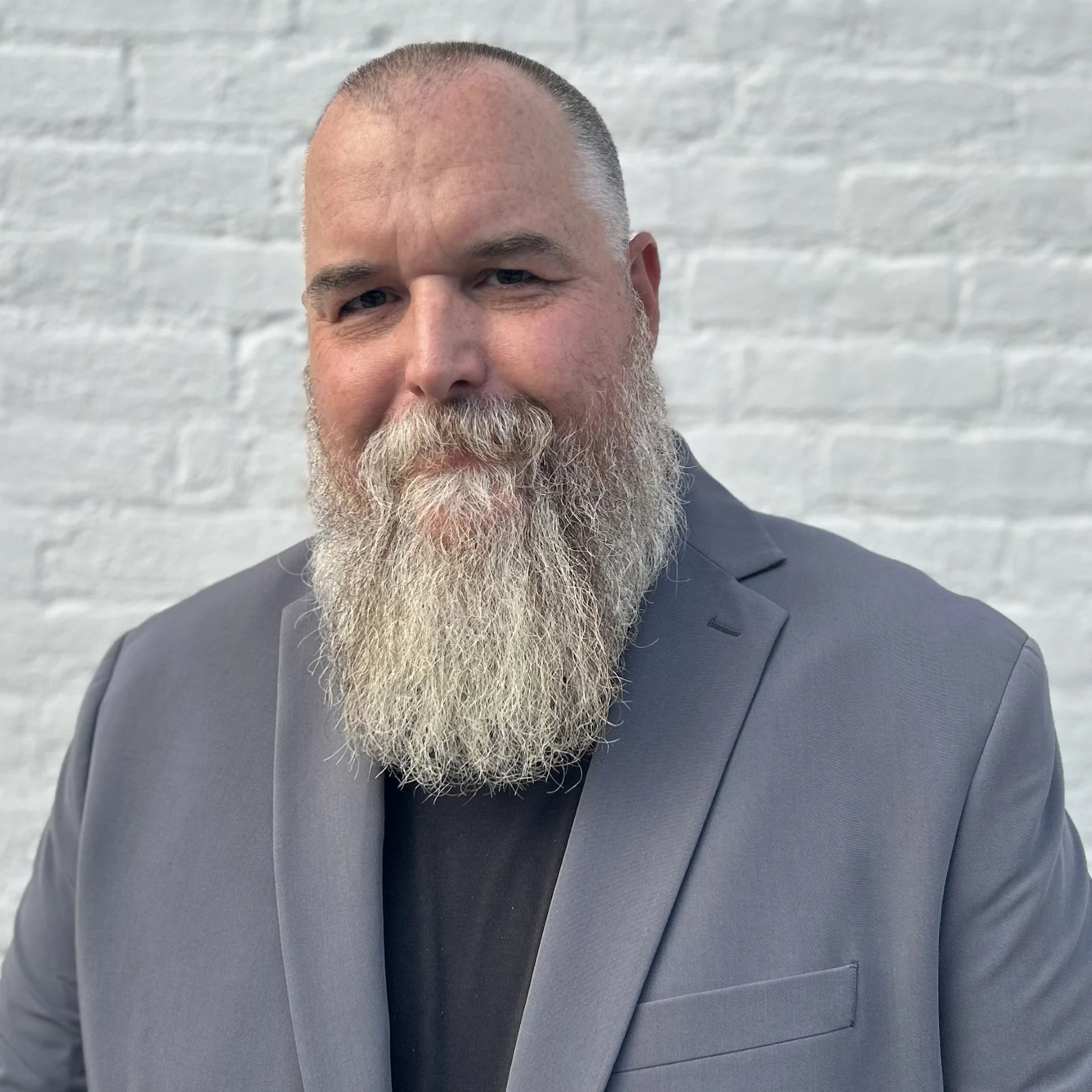 A man with a large, gray beard and short hair wearing a gray suit and black shirt, standing in front of a white brick wall.
