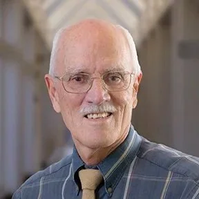 Portrait of an elderly man with white hair, glasses, wearing a plaid shirt and a tie, smiling at the camera.