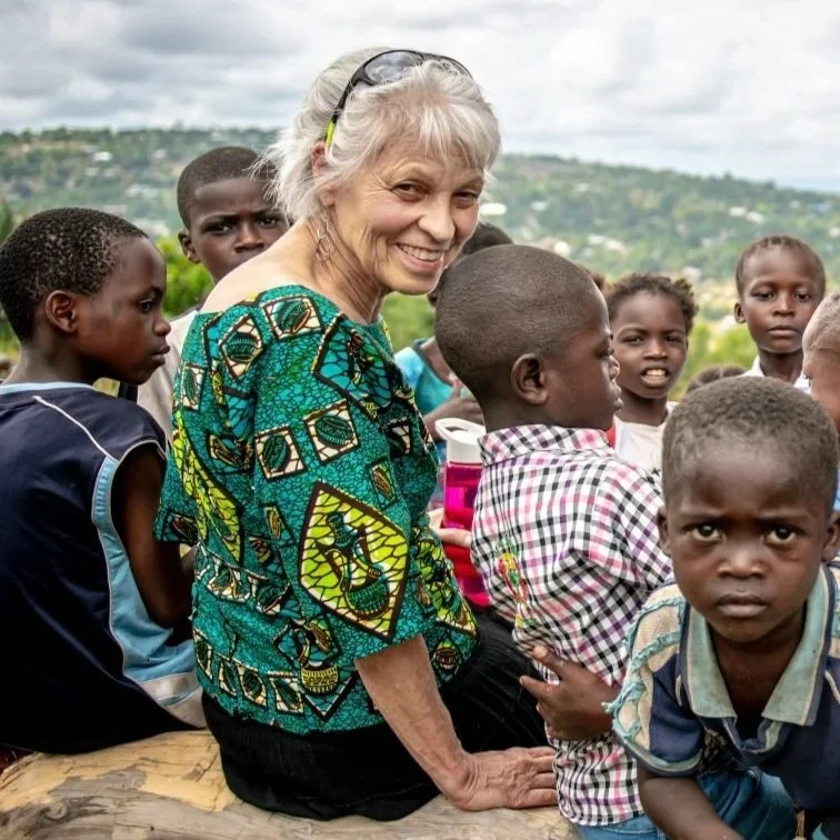An elderly woman with gray hair and glasses on her head surrounded by young children outdoors with a hilly landscape in the background.