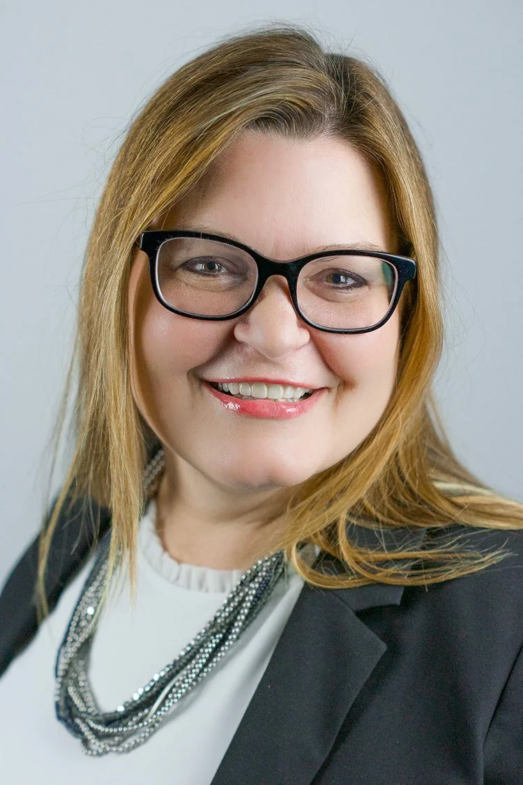 A woman with shoulder-length strawberry blonde hair, wearing glasses, a black blazer, and a white top with layered necklaces, smiling at the camera.
