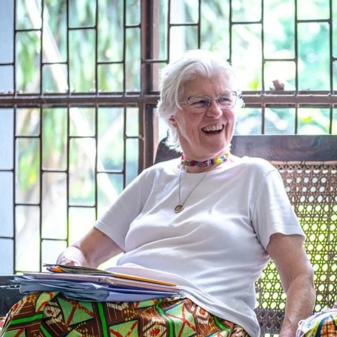An older woman with white hair, wearing glasses, a white t-shirt, and colorful patterned skirt, smiling and sitting in a room with glass windows.
