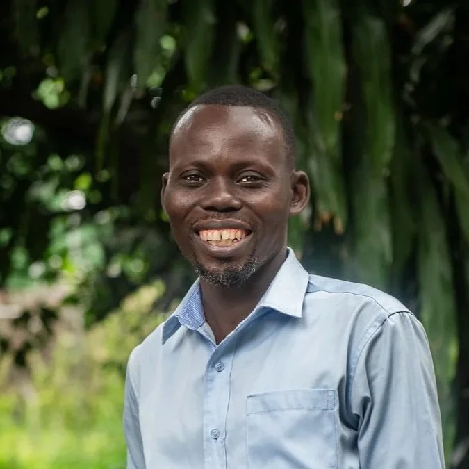 A smiling man wearing a light blue button-up shirt standing outdoors with a background of green foliage.