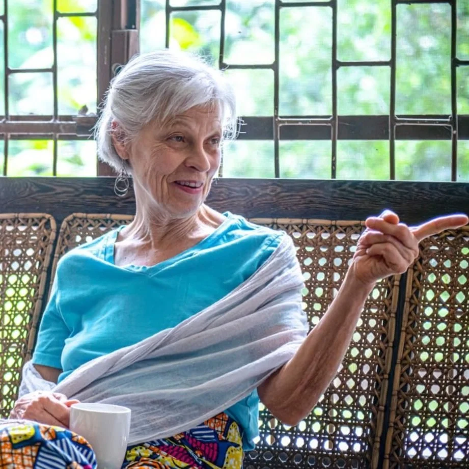An elderly woman with gray hair smiling and gesturing with her right hand, sitting inside near a window with a wooden frame and latticed glass. She is wearing a blue shirt and a white shawl, with a colorful cloth in her lap and holding a white mug.