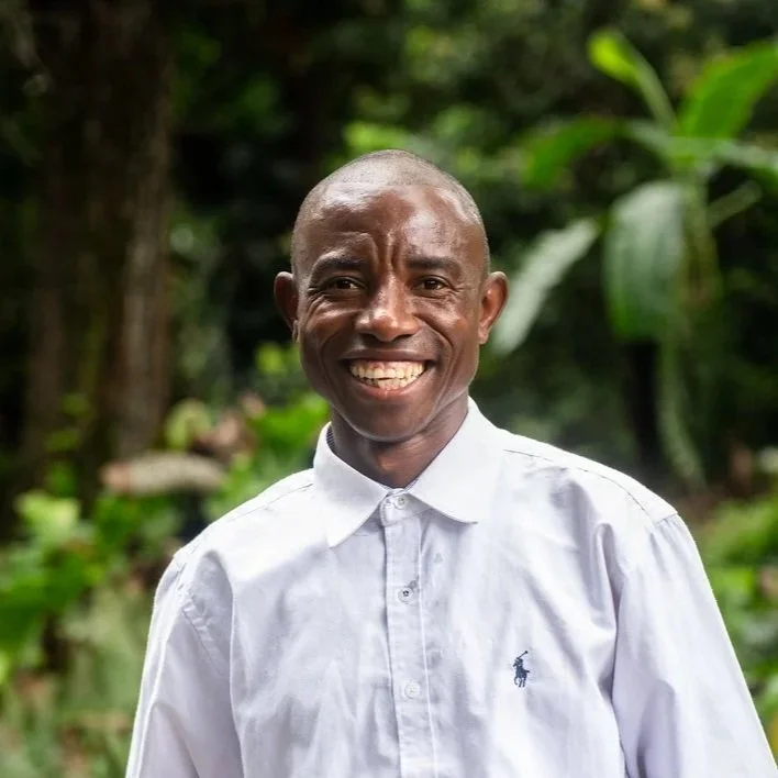 A smiling man with a shaved head wearing a white button-up shirt outdoors with green foliage in the background.