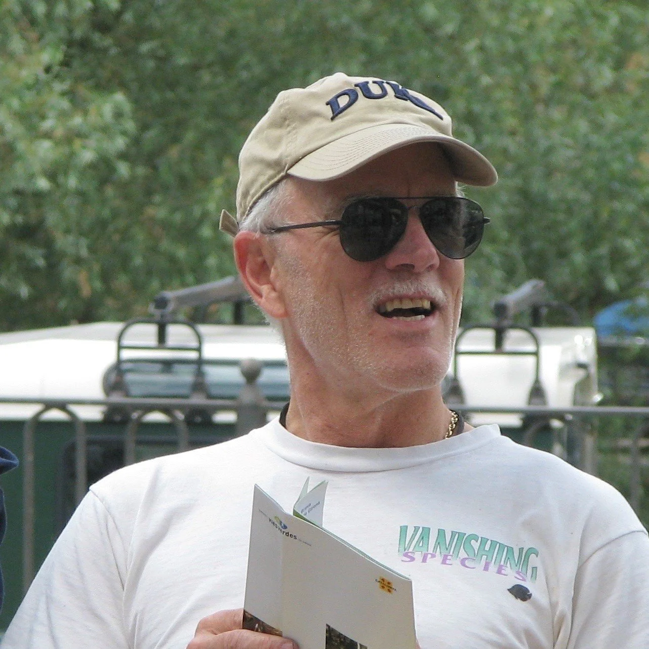 Man wearing a cap and sunglasses, holding documents outdoors.