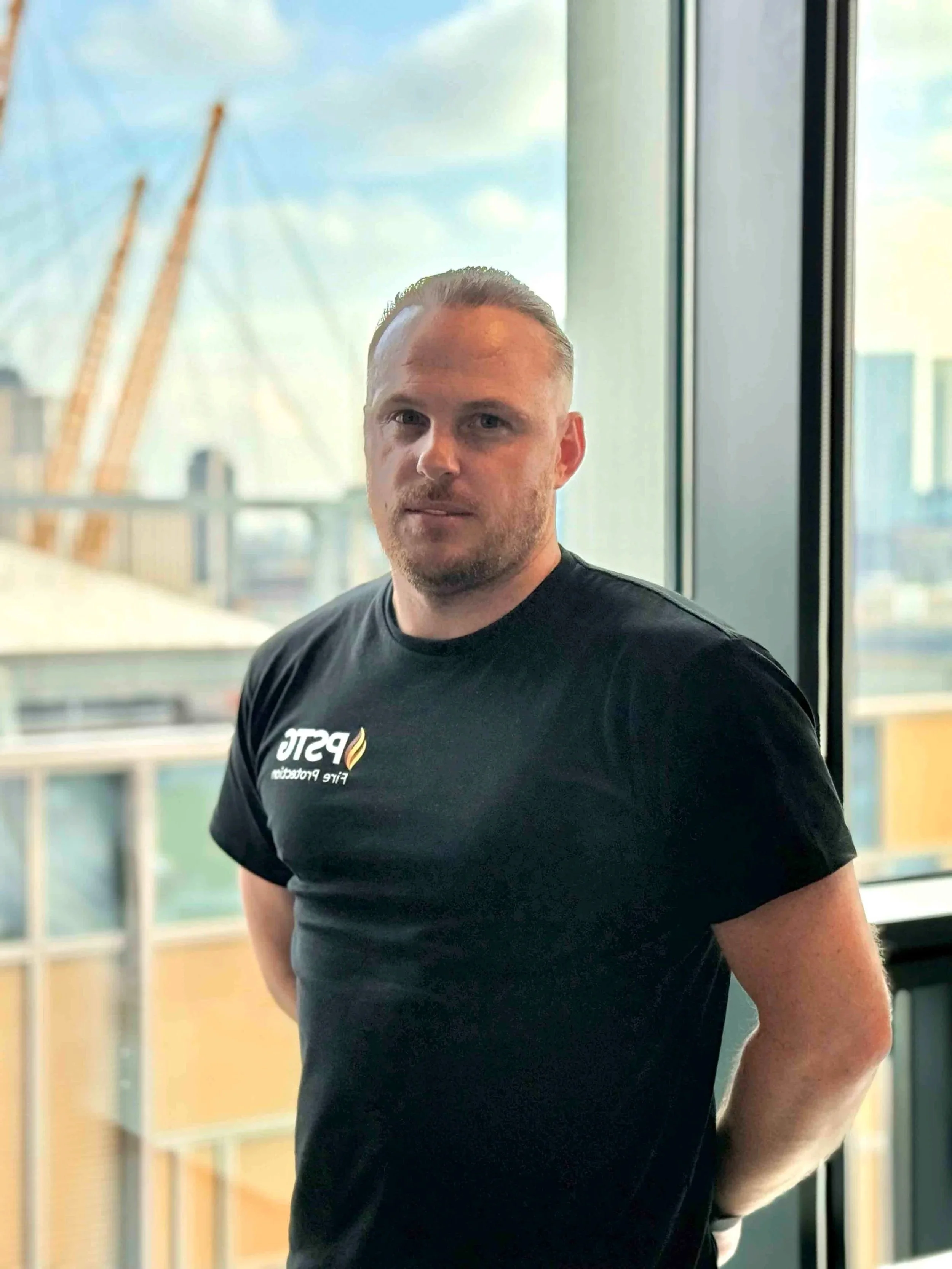 A man with short hair and a beard standing indoors near a large window, wearing a black t-shirt with a logo that says 'PSTC' and a flame symbol, with a cityscape visible in the background.
