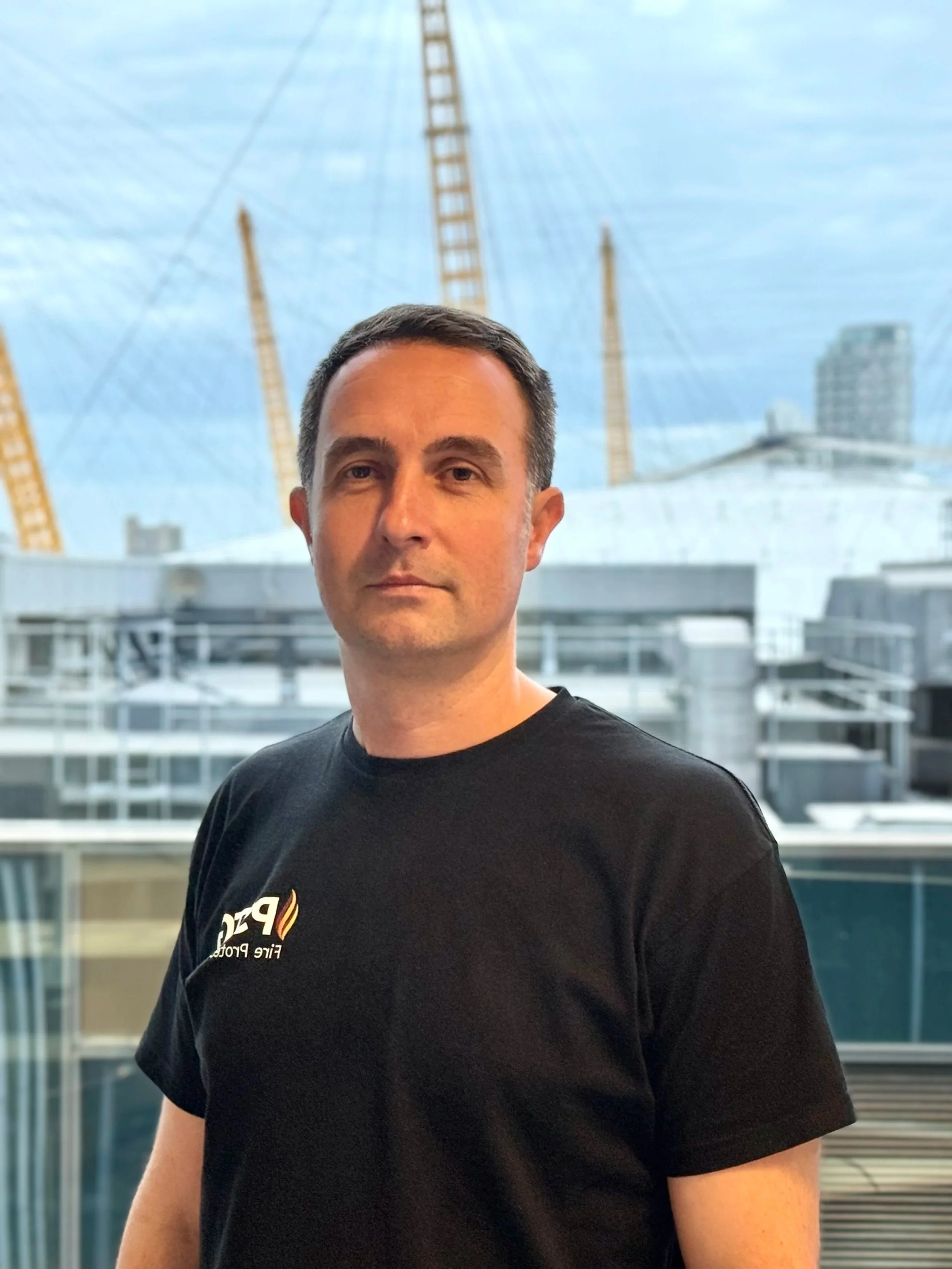 Man in black t-shirt standing indoors in front of large window with city buildings and a Ferris wheel outside.