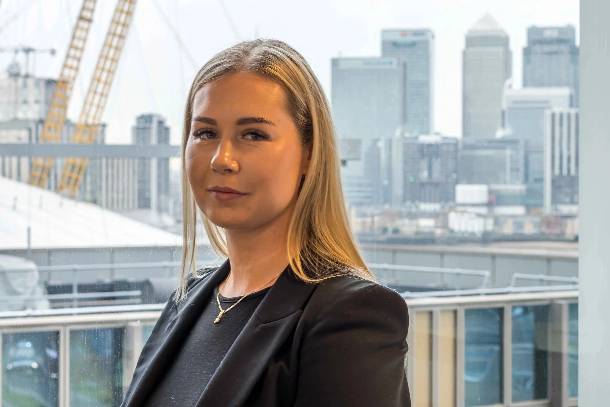 A young woman with straight blonde hair wearing a black blazer and top, standing in front of a large window with a cityscape background.