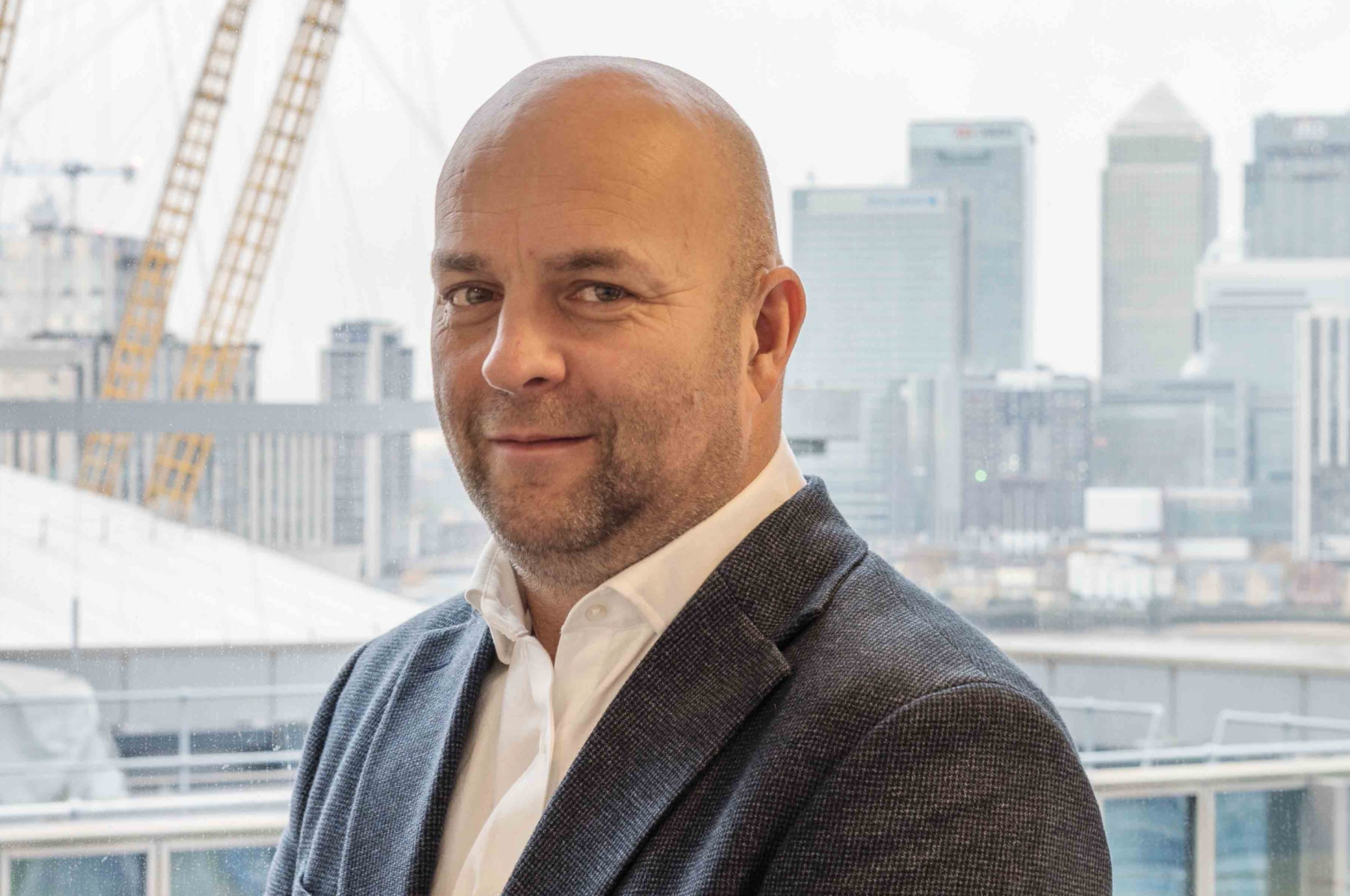 A man with a bald head and beard wearing a white shirt and dark blazer, standing in front of a cityscape with tall buildings and cranes.