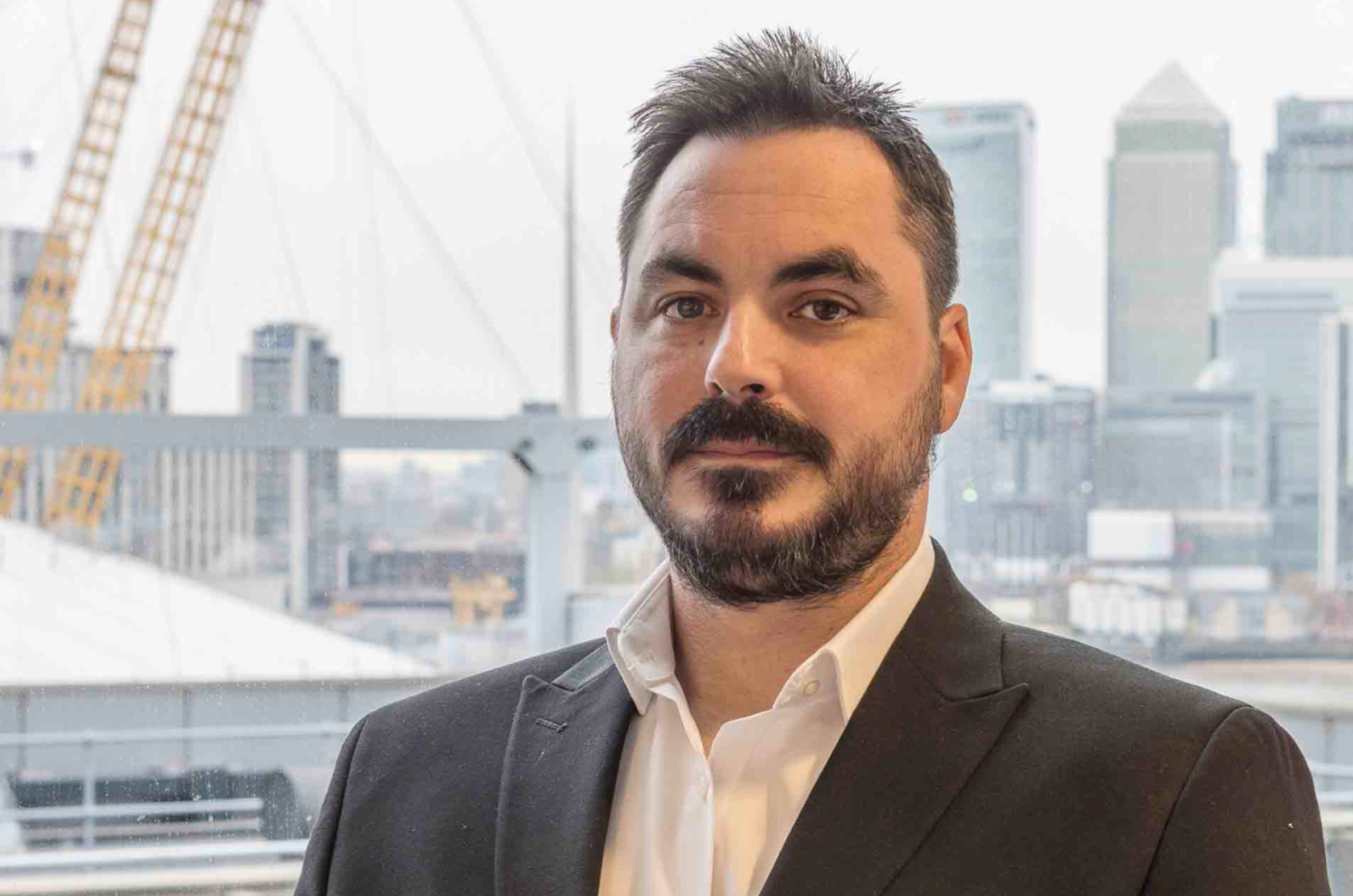 A man with dark hair, a beard, and a mustache wearing a black suit and white shirt, standing indoors with a city skyline backdrop, including buildings and construction cranes.