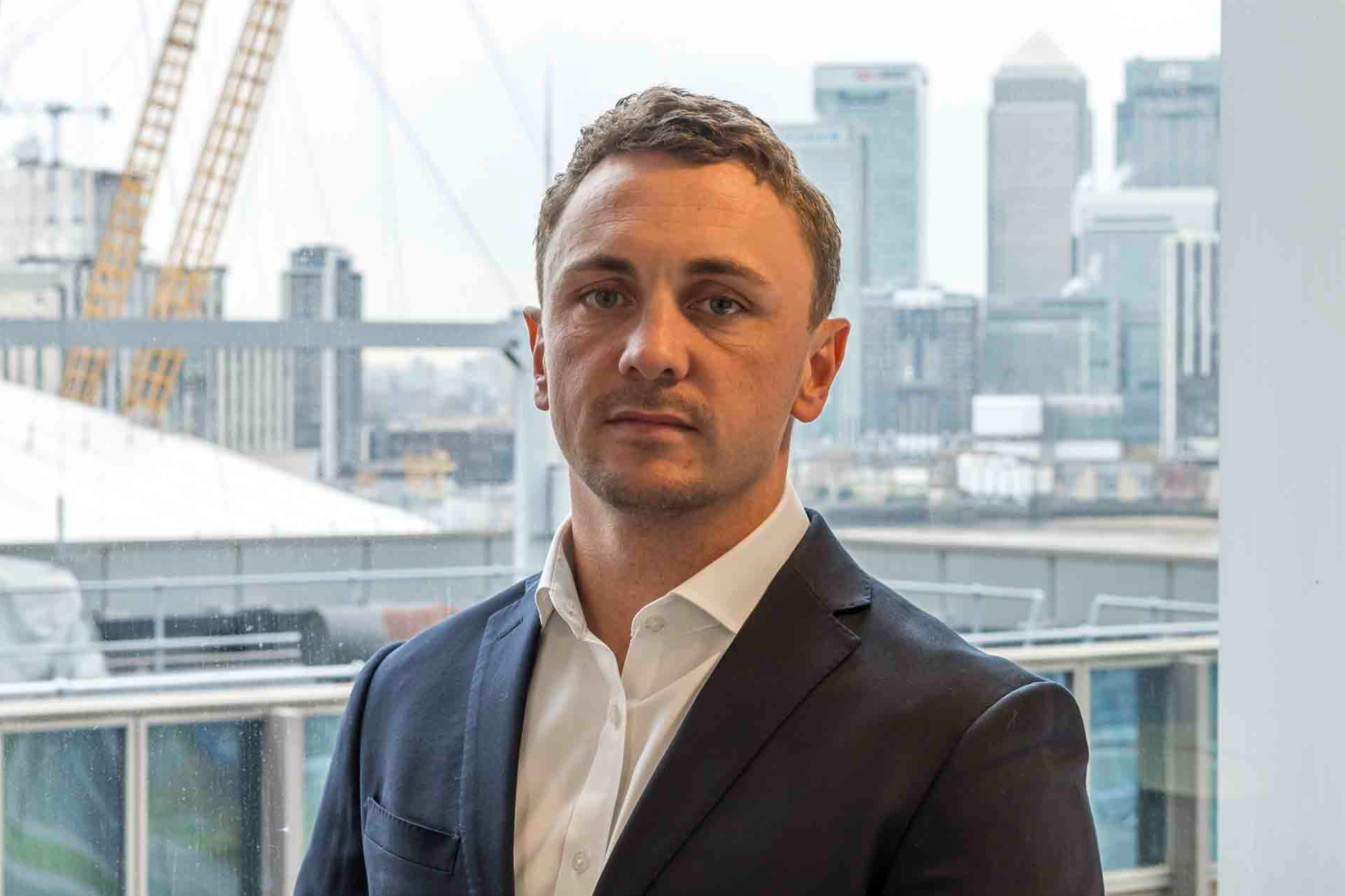 A man in a suit with short brown hair and a serious expression, standing in front of a window with a cityscape in the background.