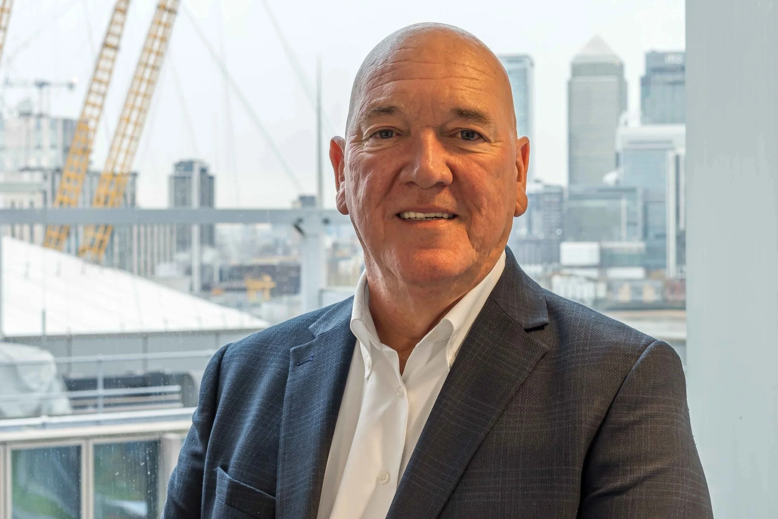 Headshot of a middle-aged man in a dark suit and white shirt, smiling, with a city skyline and bridge visible in the background.