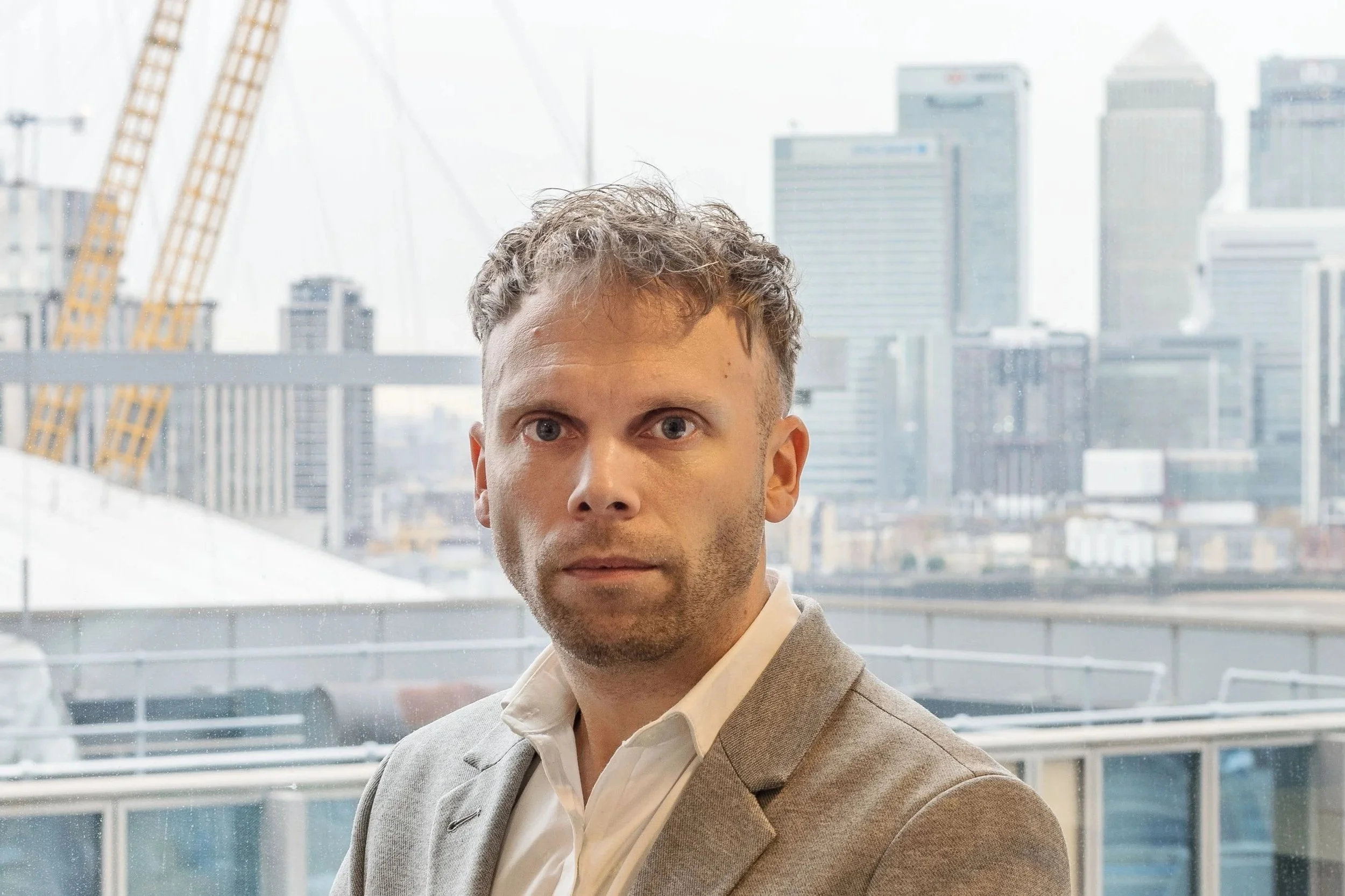 A man in a gray suit and white shirt standing inside a building with a city skyline and construction cranes in the background.