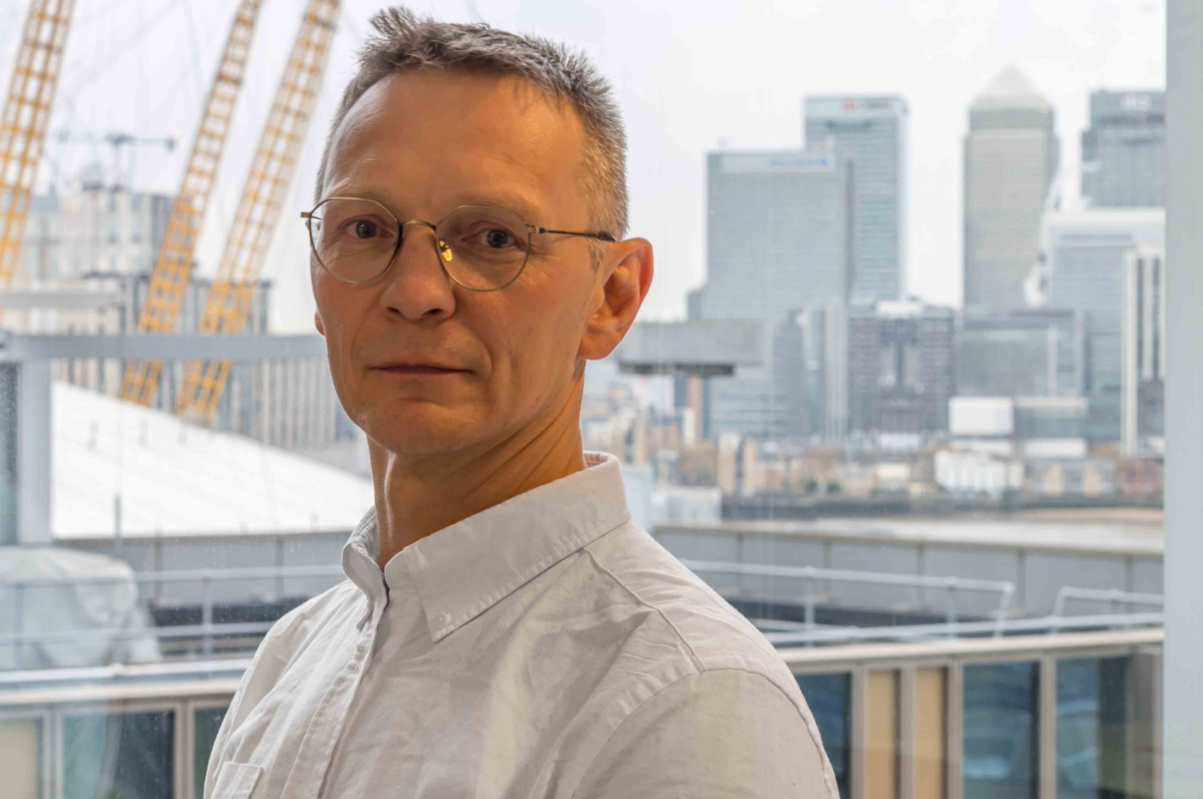 A middle-aged man with glasses and short gray hair standing indoors with a city skyline visible through a large window behind him.