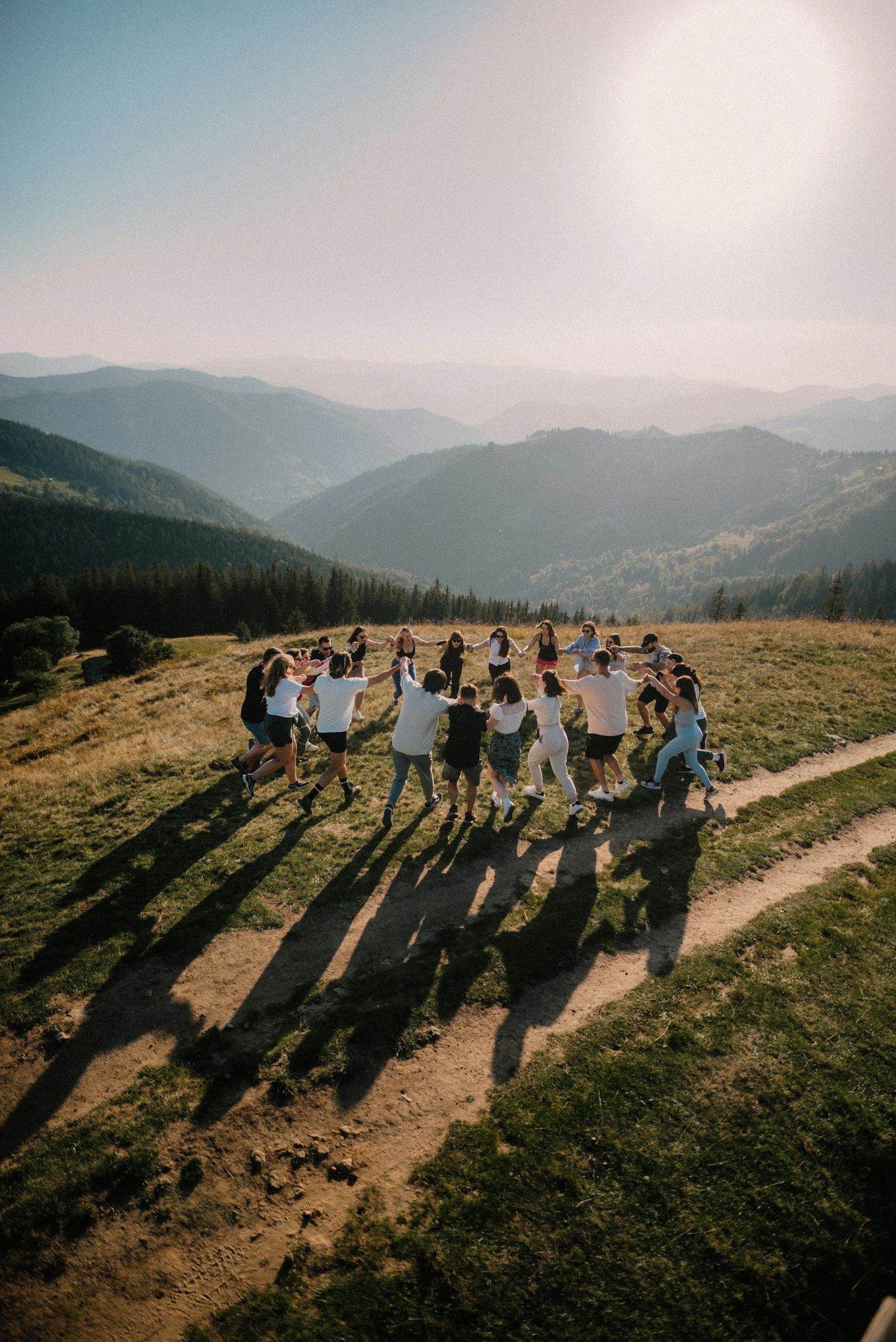 People holding hands and dancing in a circle on a grassy hilltop with mountain range in the background, during late afternoon or early evening.