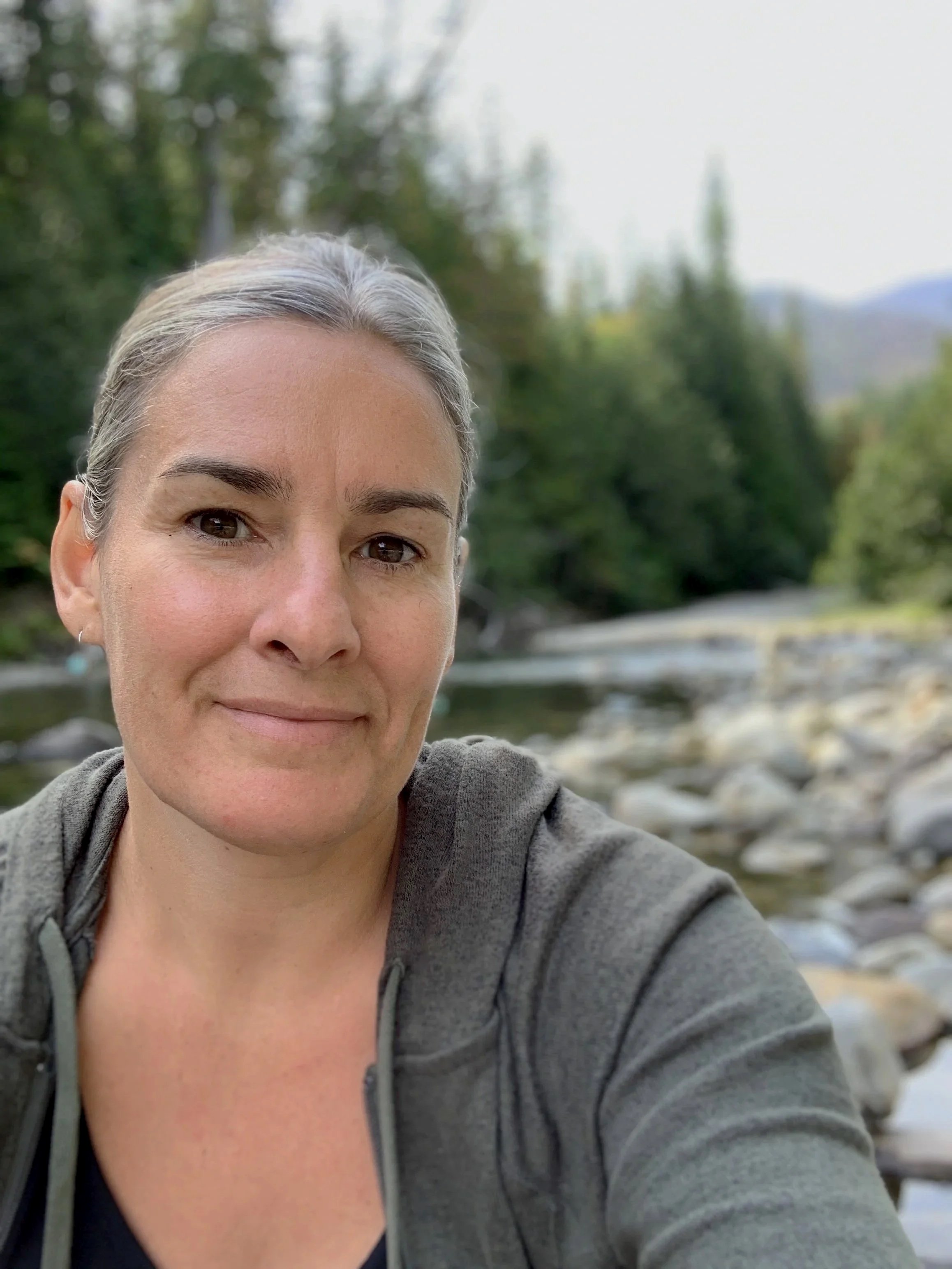 Close-up of a woman with gray hair and brown eyes smiling outdoors near a river with a rocky shoreline and green trees in the background.