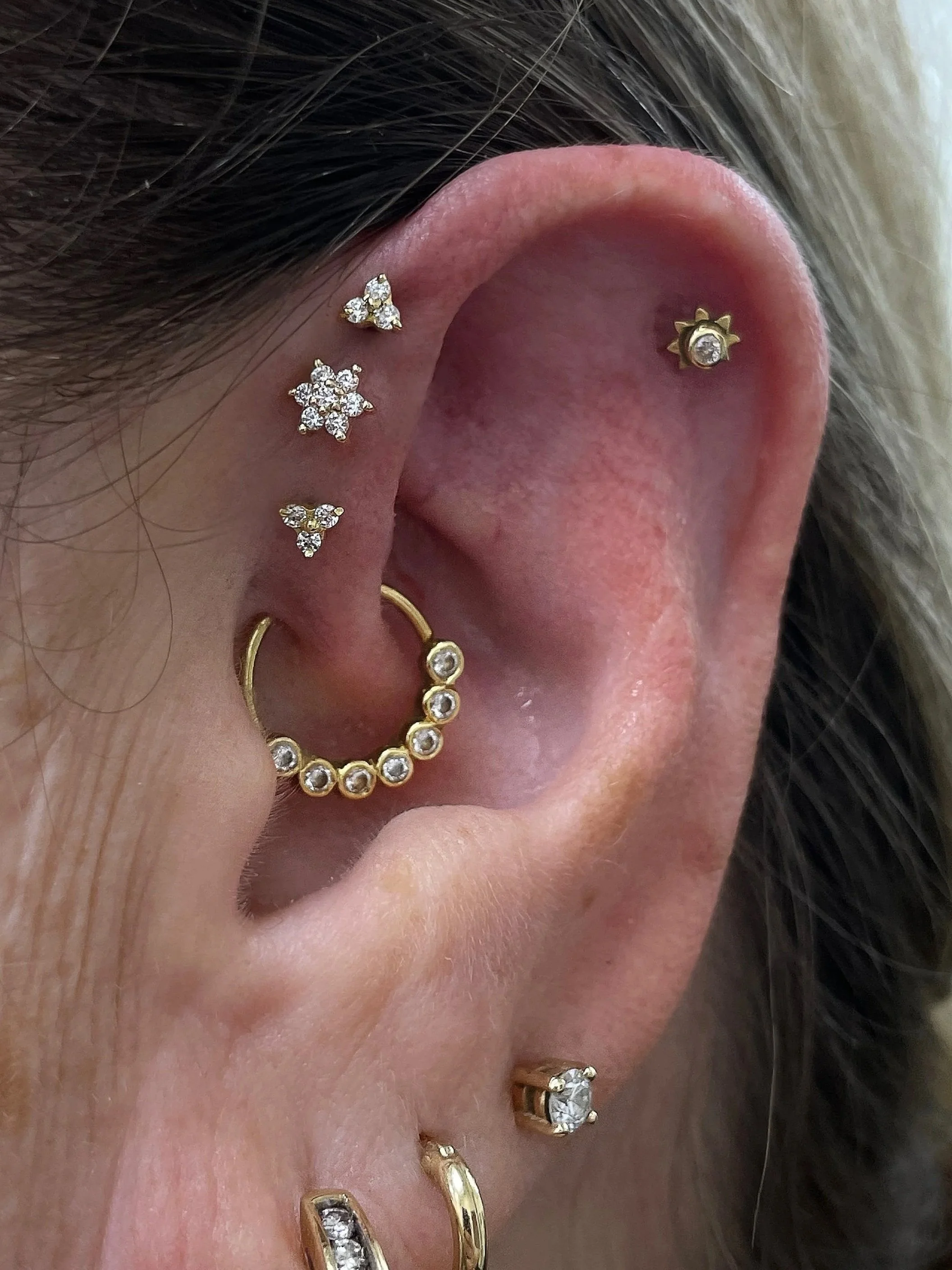 Close-up of a human ear with multiple gold earrings, including a small flower-shaped cluster with clear stones, a tiny sun-shaped stud, a hoop with clear stones, and several other small earrings with clear stones.