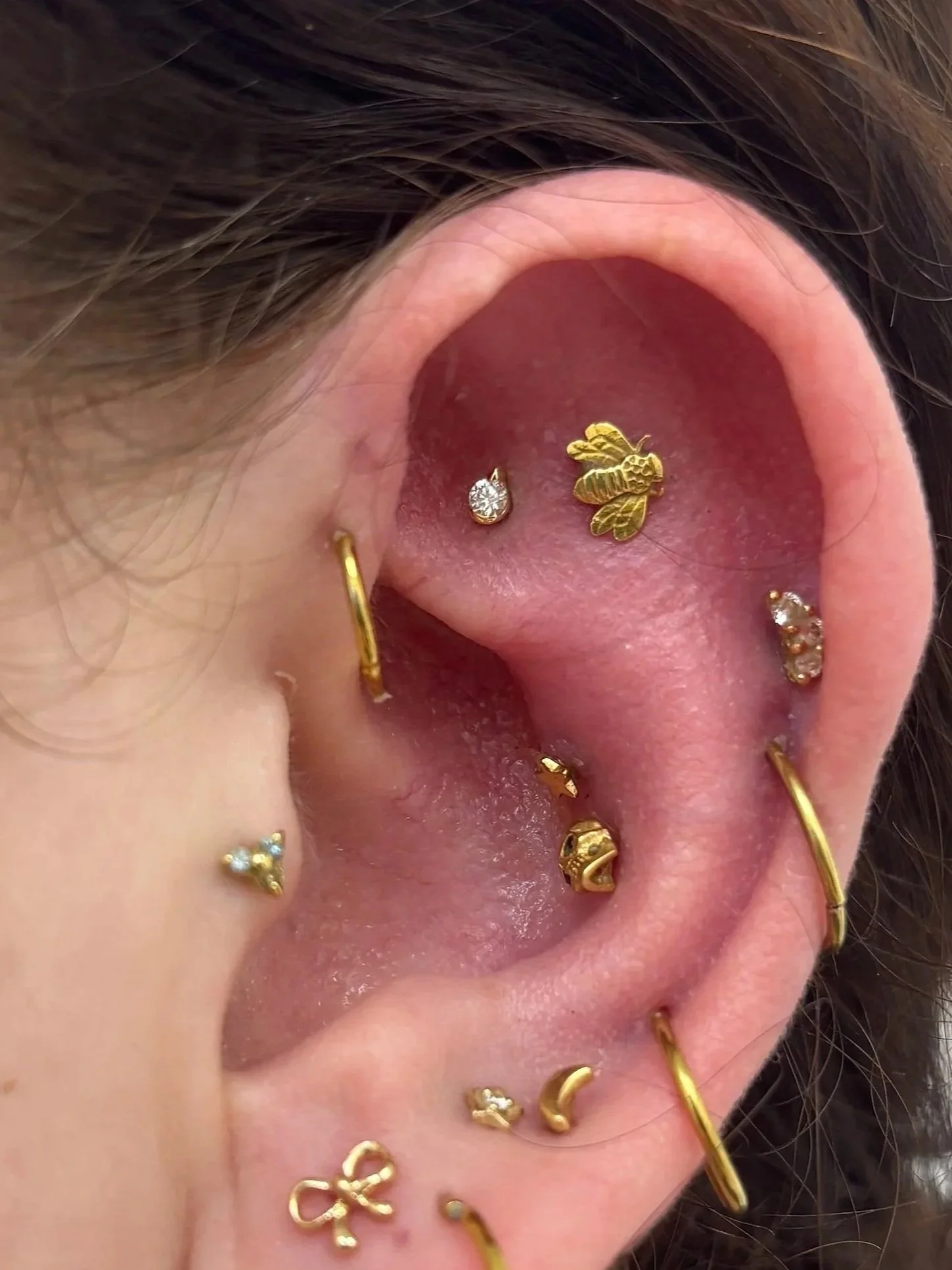 Close-up of a person's ear with multiple gold earrings, including hoops, studs, and decorative designs, with some jewelry featuring small gemstones.