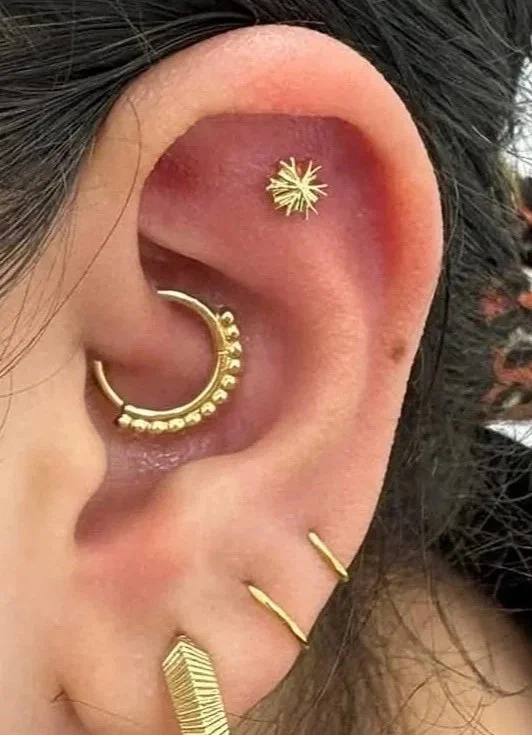 Close-up of a person's ear with multiple gold earrings, including a star-shaped stud, a hoop with beads, a small double bar, and a textured rectangular piece.
