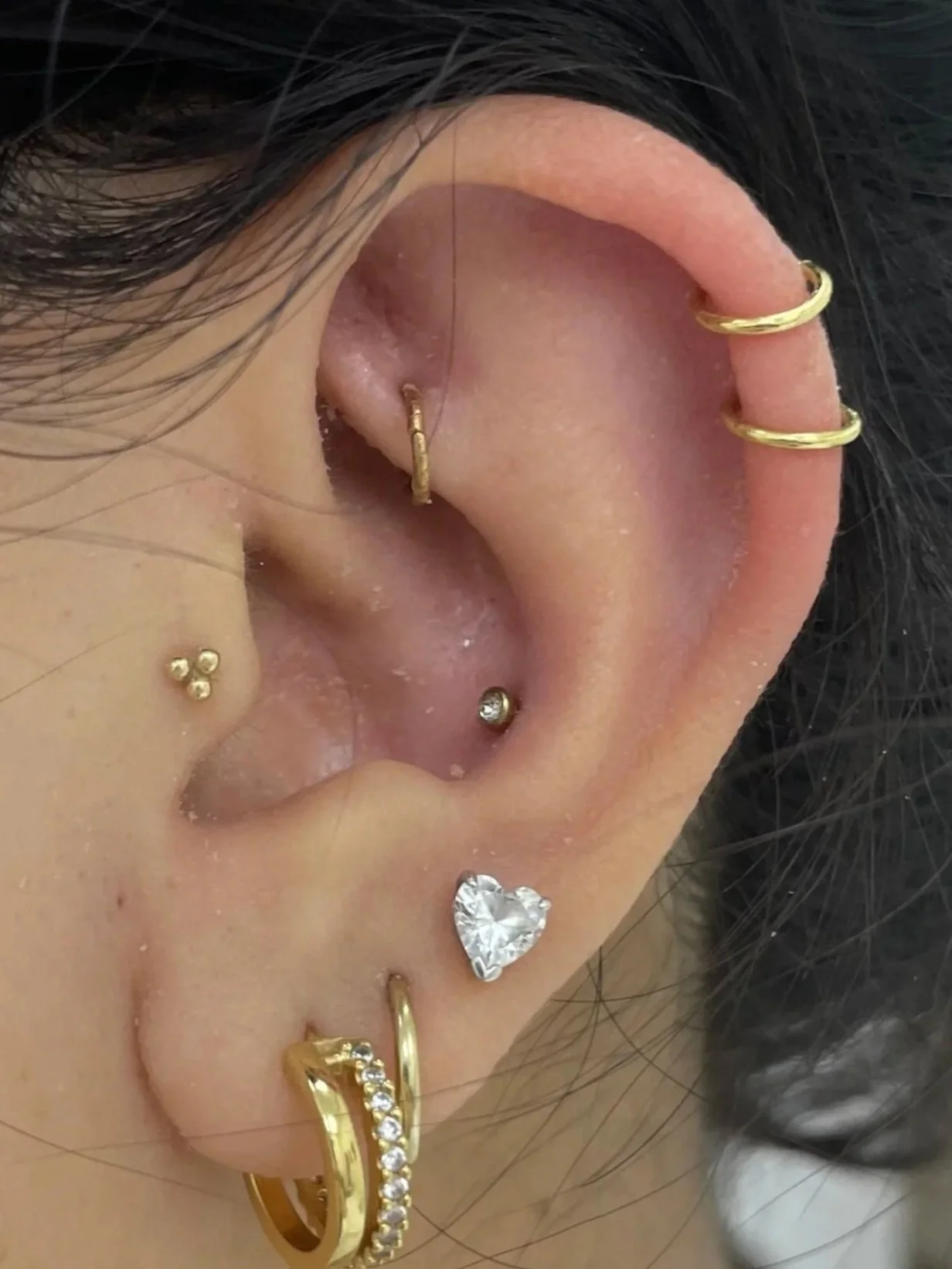 Close-up of a person's ear with multiple gold and silver earrings, including hoops, studs, and a heart-shaped dangle, with black hair visible.