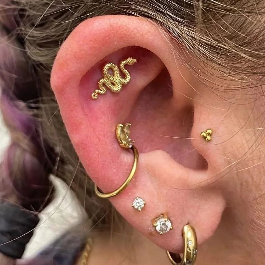 Close-up of a person's ear adorned with multiple gold earrings, including a script word earring, a hoop with a cuff, a small ball stud, a diamond stud, a cluster diamond stud, and a gold hoop with embedded stones.