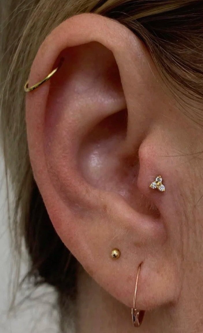 Close-up of a person's ear with multiple piercings: a gold hoop on the helix, a gold stud on the lobe, a small triangle-shaped cluster of three gemstones on the tragus, and a thin gold hoop on the lower lobe.