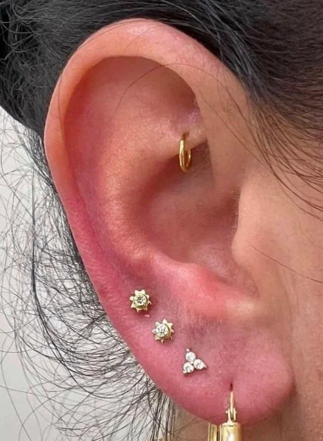 Close-up of a woman's ear with multiple earrings, including a small gold hoop on the upper cartilage, two stud earrings with a sun-like design and a clear central stone, and a gold hoop earring at the earlobe.