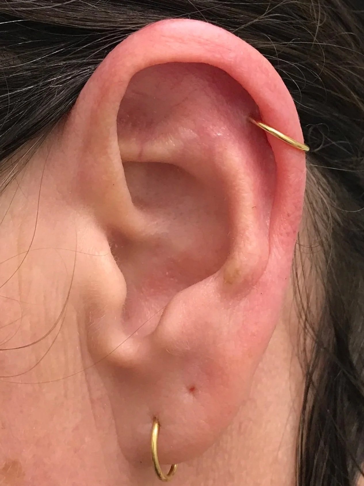Close-up of a person's ear with a gold hoop earring in the earlobe and a small gold hoop in the upper cartilage, with visible hair and skin details.