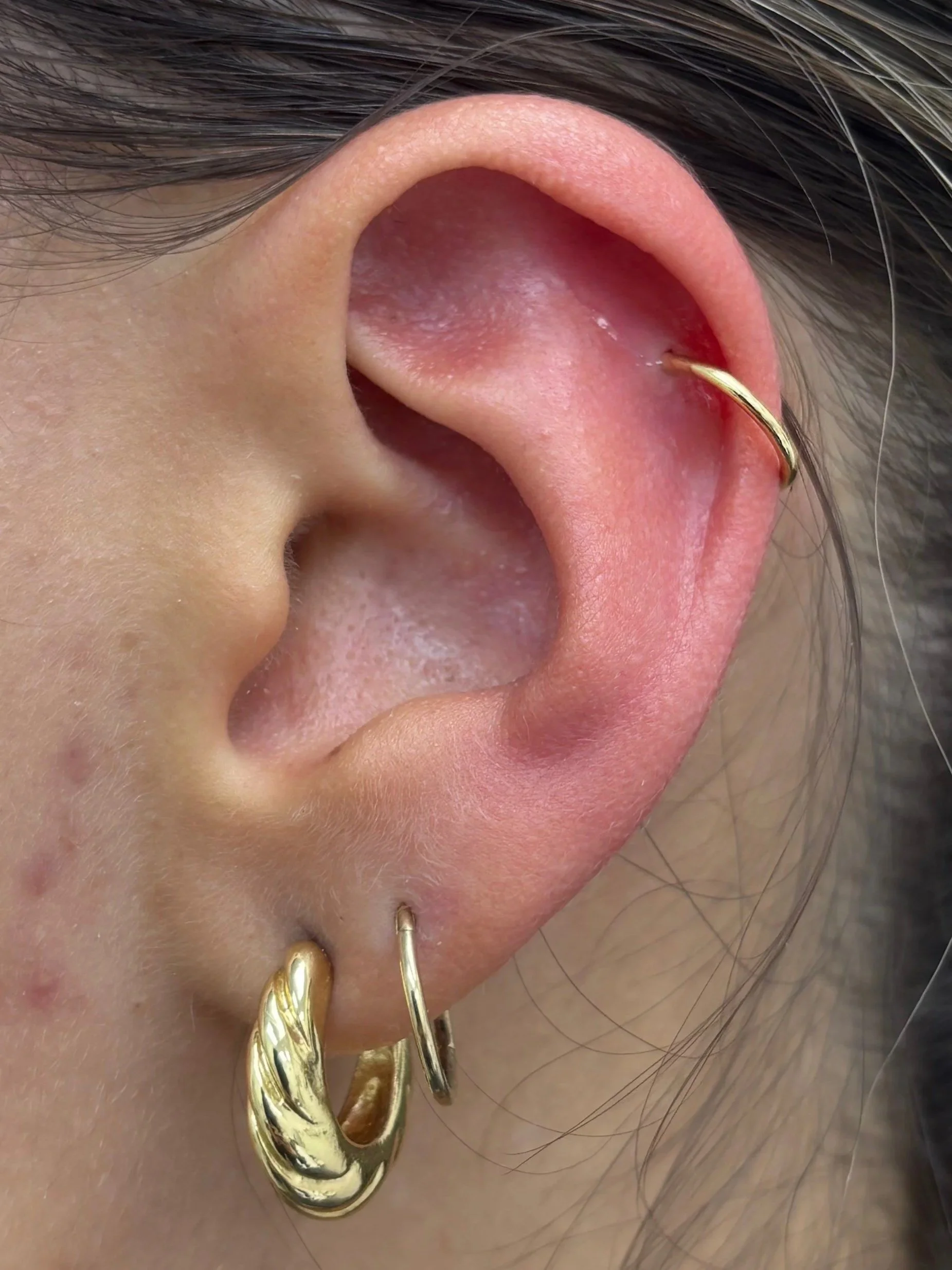 Close-up of a person's ear with multiple gold earrings, including a hoop, a twisted hoop, and a tiny hoop, with hair and skin visible.