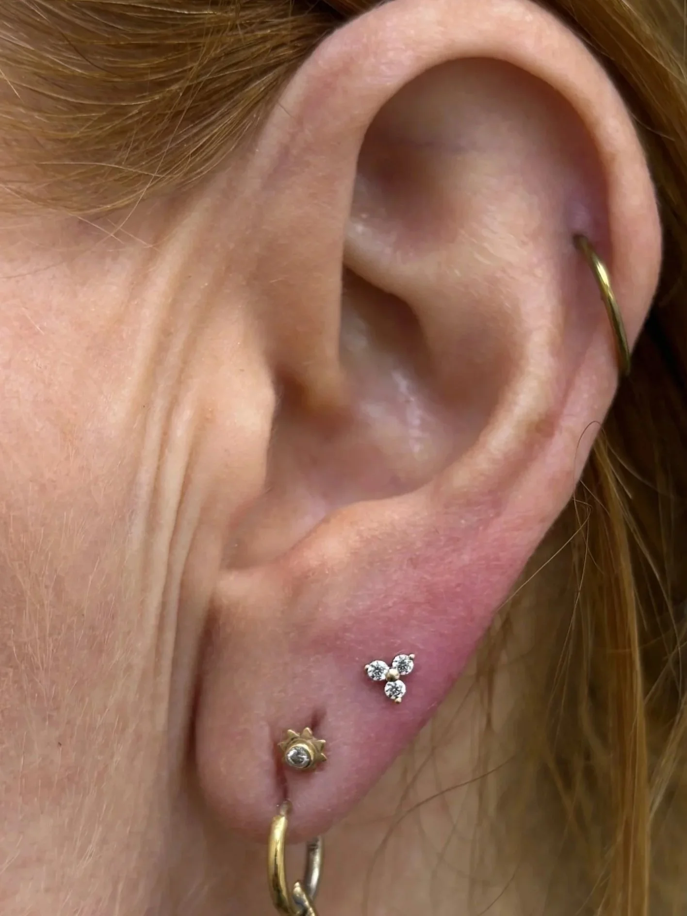 Close-up of a person's ear with multiple earrings: a gold hoop at the earlobe, a small star-shaped ring with a gemstone, a cluster of diamond-like gemstones, and a small gold hoop with a gemstone.