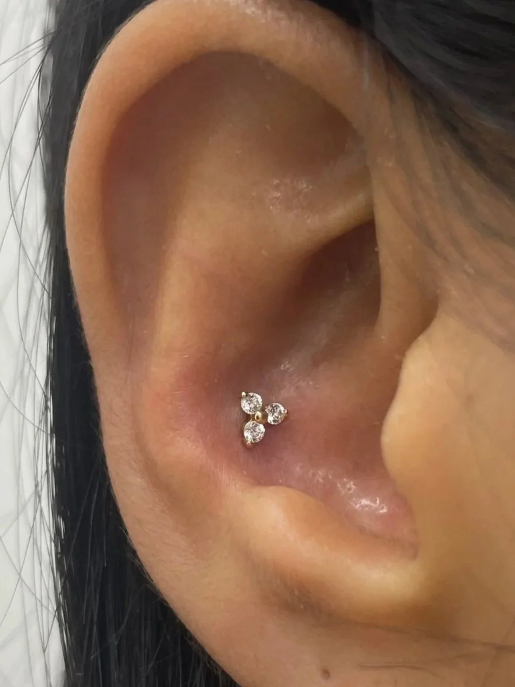 A close-up of a woman's ear with a triple diamond cartilage earring and a gold hoop earring, black hair visible around the ear.