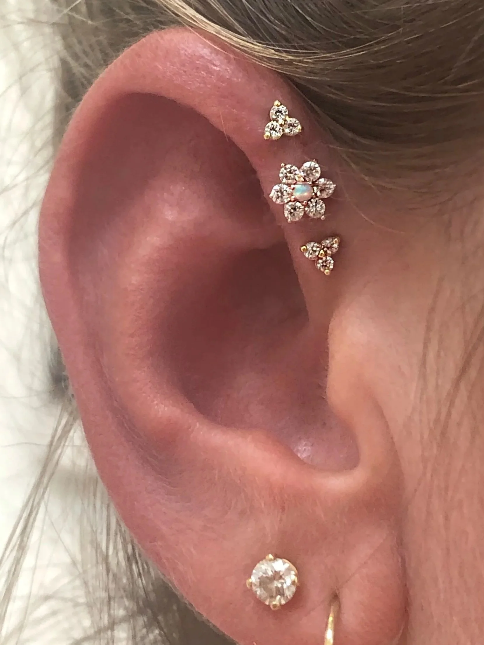 Close-up of a person's ear with multiple earrings, including a gold hoop, a round diamond stud, and three small earrings with butterfly, flower, and butterfly designs adorned with small gemstones.