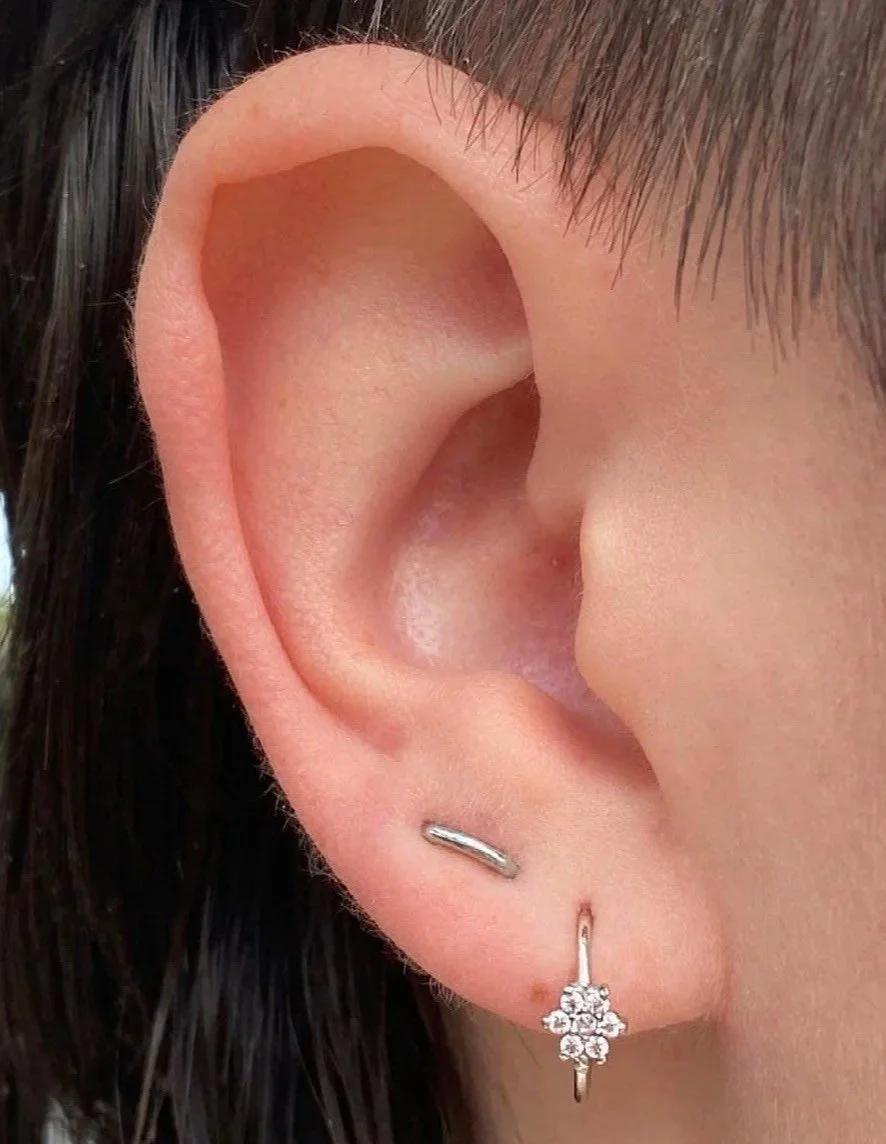 Close-up of a person's ear with two earrings: a small silver hoop in the upper cartilage and a dangling earring with a floral cluster of small diamonds.