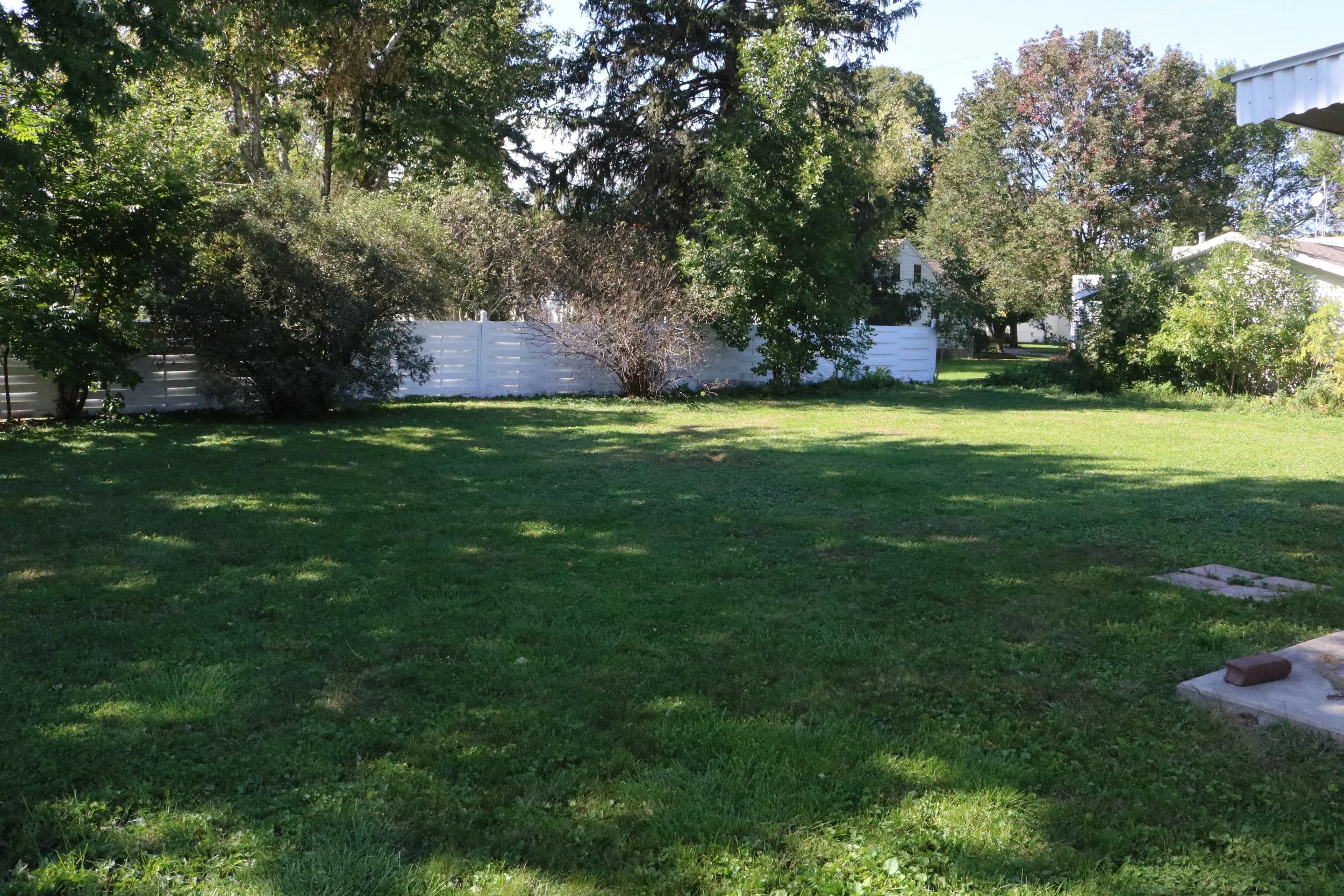 A backyard with green grass, surrounded by trees and a white wooden fence.