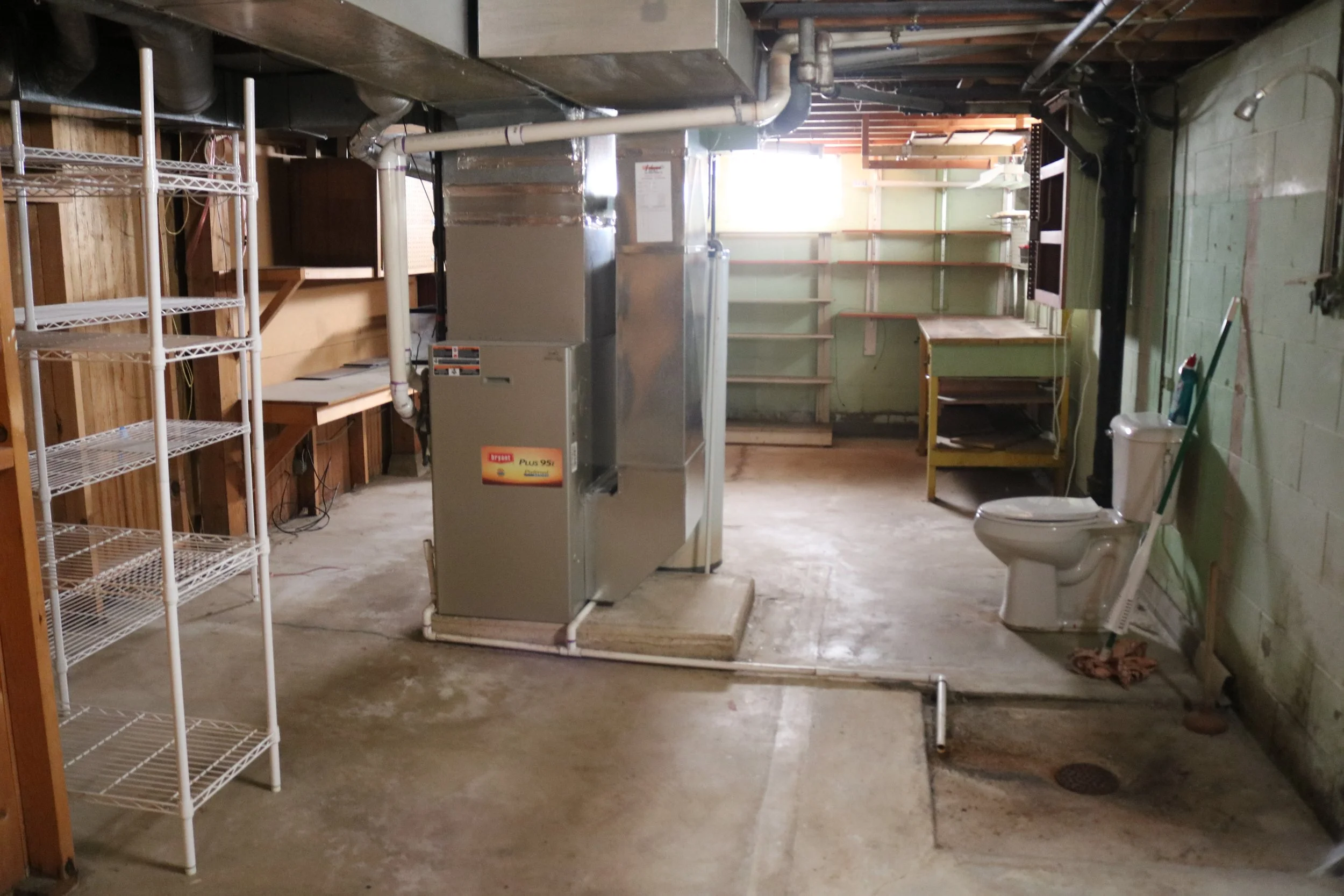 Unfinished basement with HVAC unit, white wire shelving, workbenches, toilet, and utility sink.