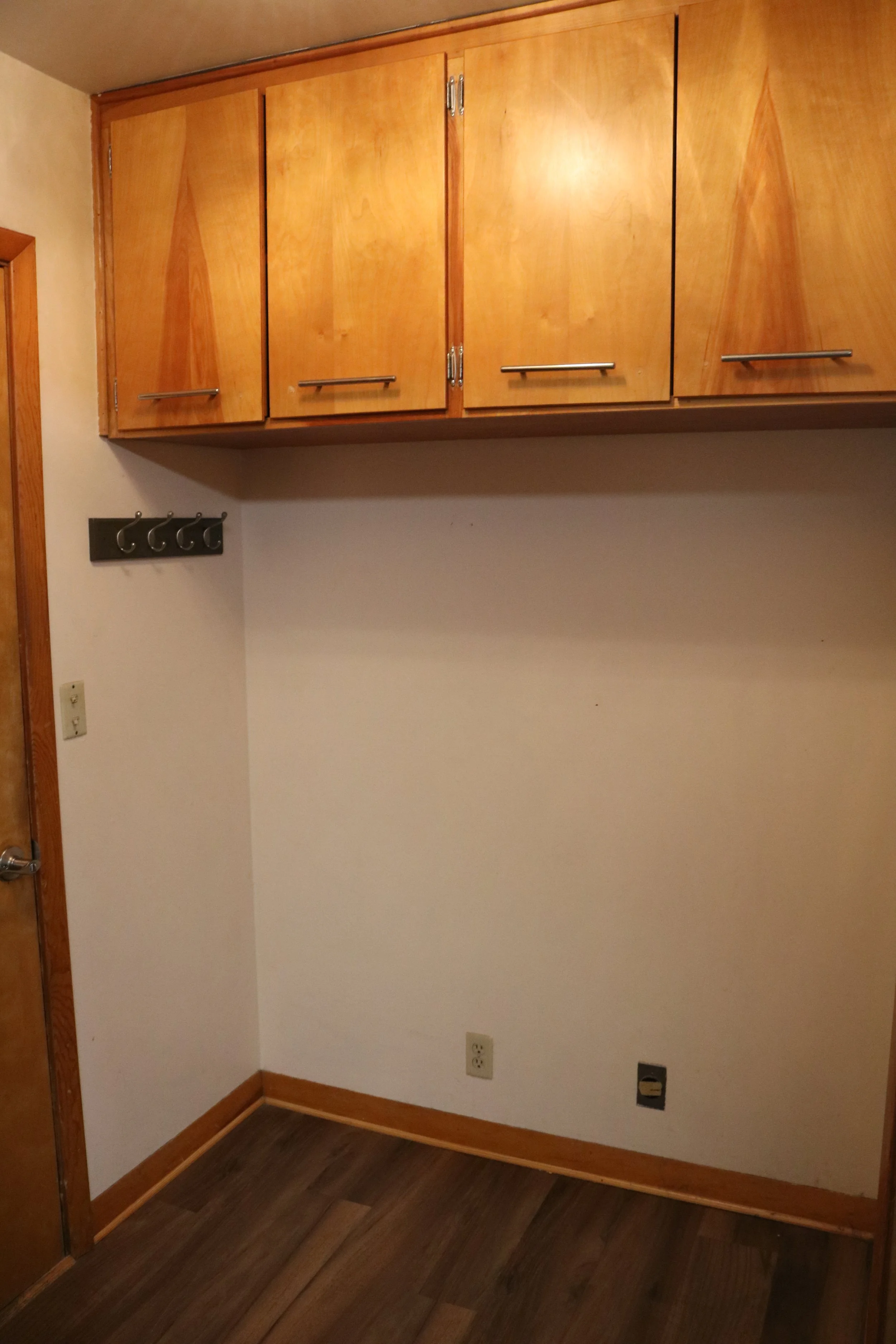 Wooden cabinets above an empty space with wall hooks on the left, located in a room with wooden flooring and a white wall.