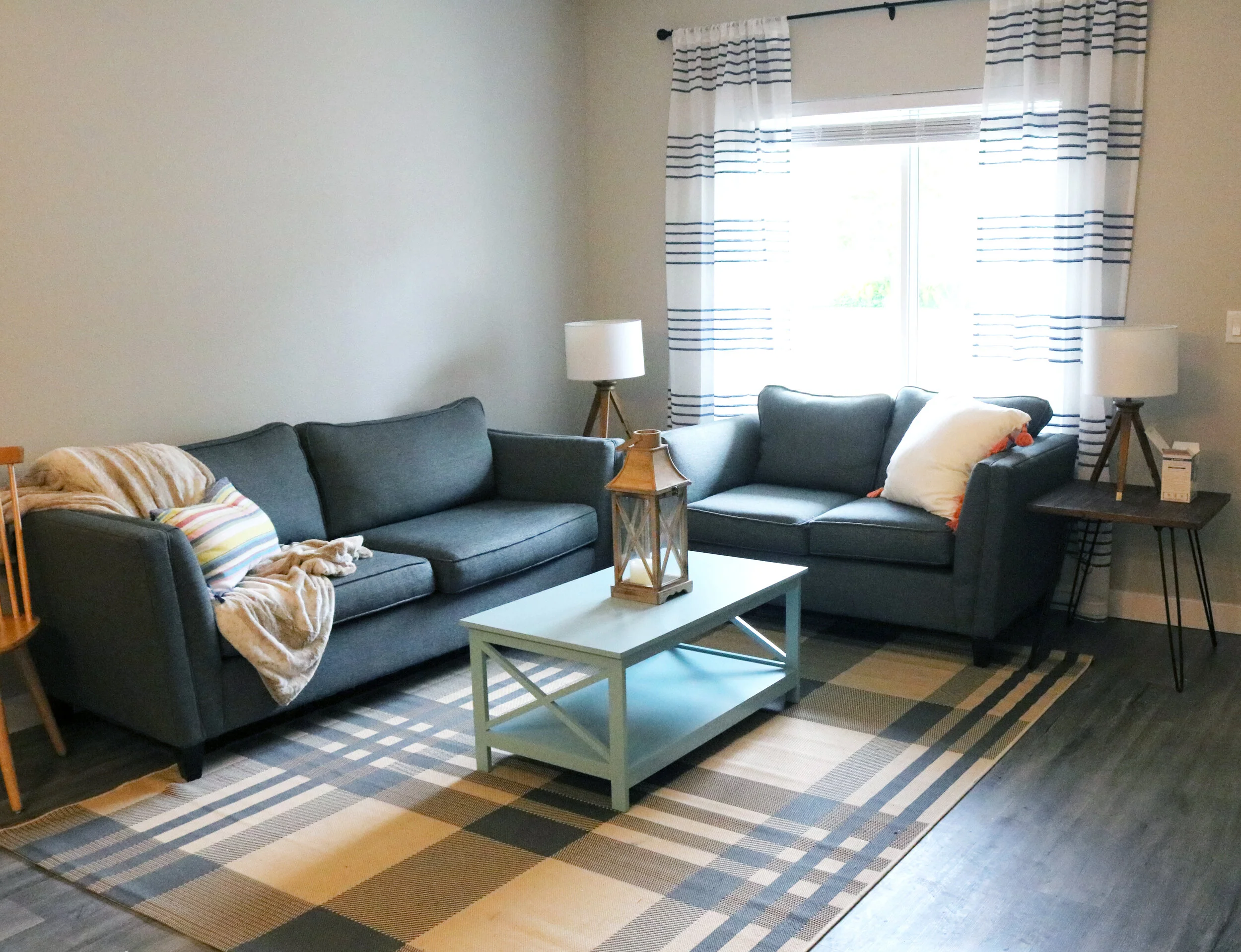 Cozy living room with gray sofas, patterned rug, and decorative pillows. A coffee table with a lantern is centered, flanked by table lamps beside the couches. Natural light filters through striped curtains.