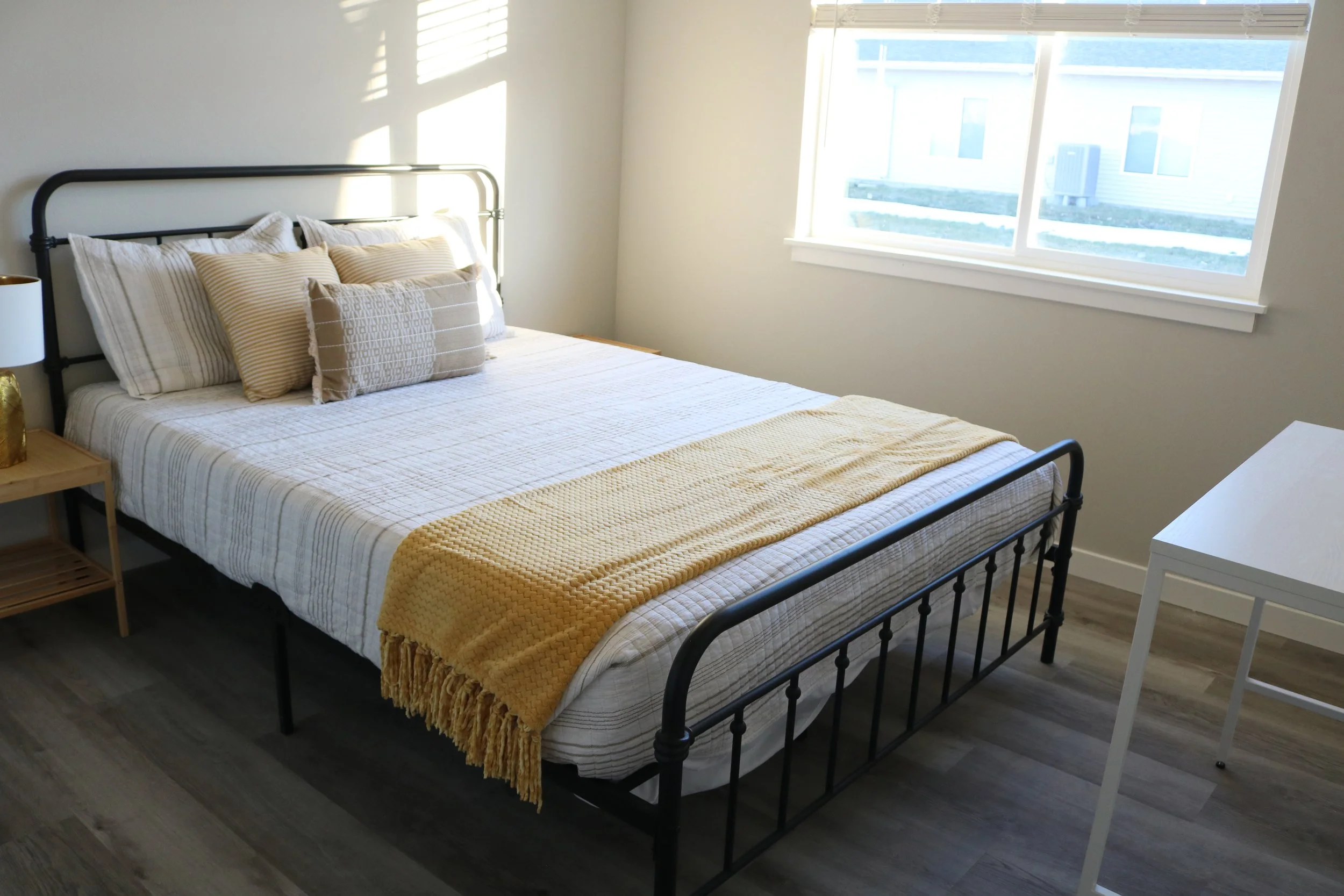 Minimalist bedroom with a metal bed frame, white and beige bedding, yellow throw blanket, side table, and a window with blinds.