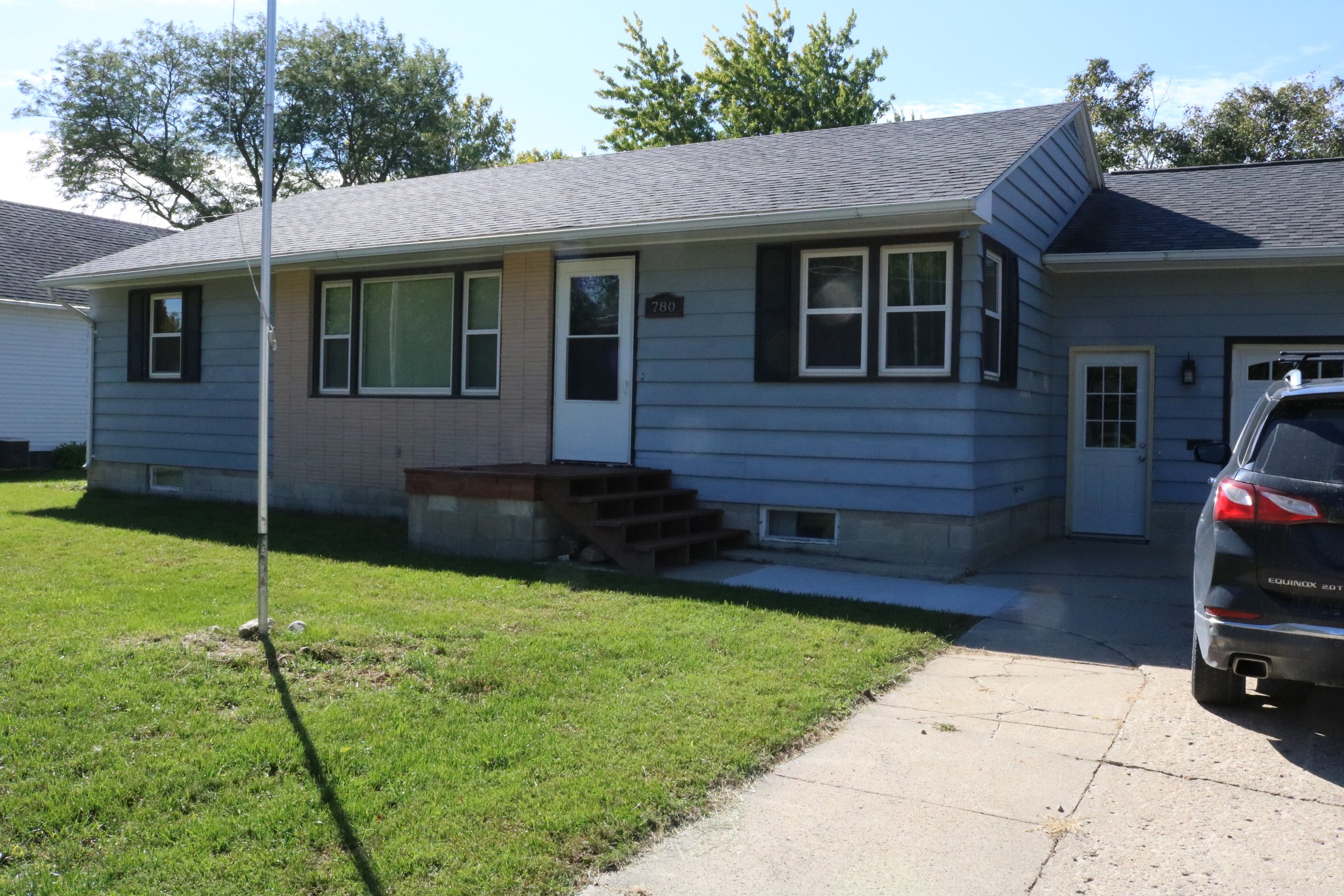 Single-story house with blue siding, a front door with small steps, a grassy lawn, and part of a car visible in the driveway.